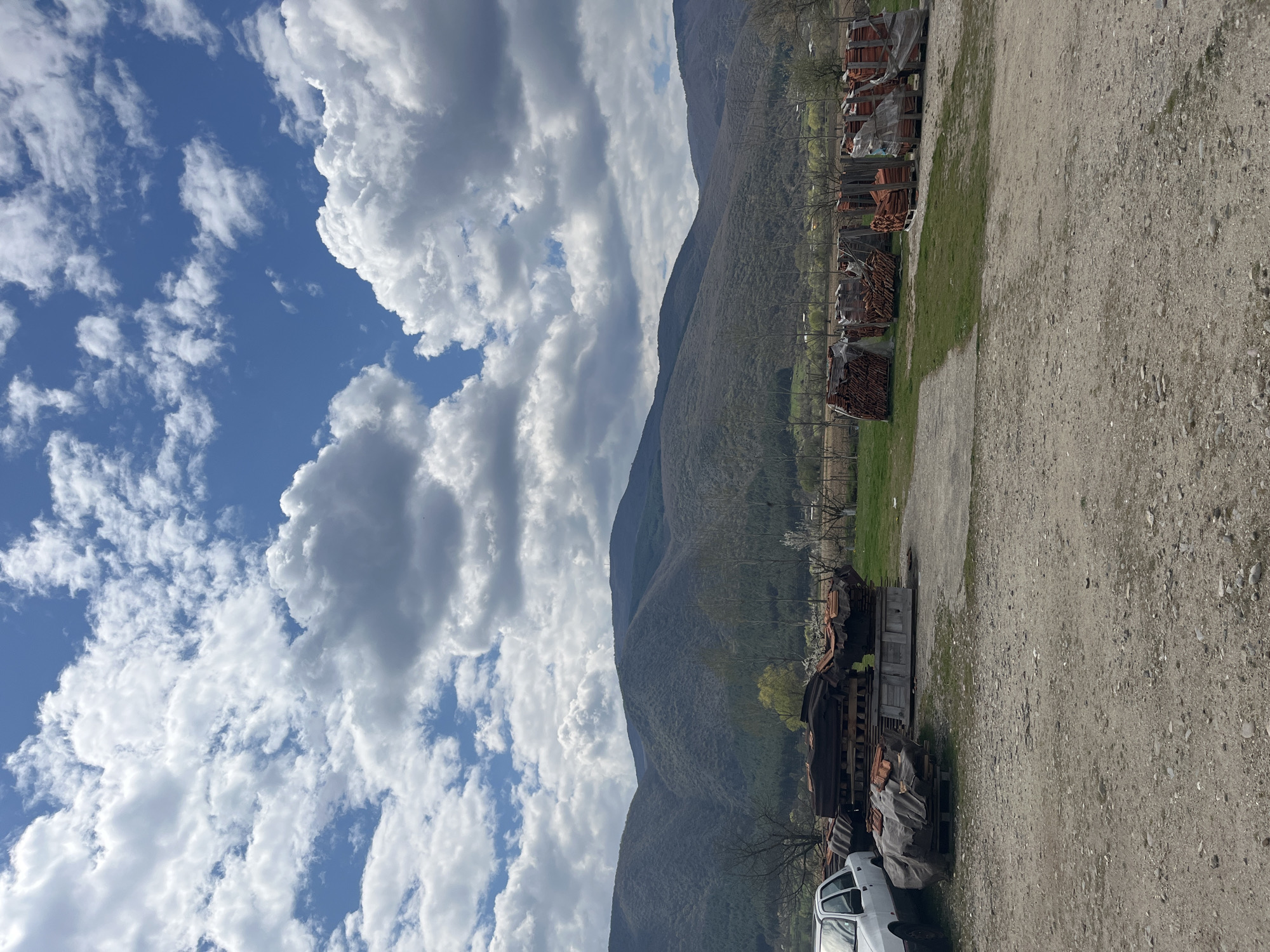 The image shows a landscape with a mountain range stretching across the center. To the left is a blue sky filled with white, puffy clouds. To the right of the mountains, there is a barren, dirt-covered area. Rows of stacked, terracotta-colored materials, possibly roof tiles or bricks, line the boundary between the dirt area and the mountains. A few trees are visible, sparsely dotting the landscape. A white car is partially visible in the lower-right corner. The image is somewhat rotated, and the specific location in Goce Delcev, Bulgaria, cannot be definitively determined from this image alone. The scene appears to be an outdoor storage or work area at the foot of a mountain range.