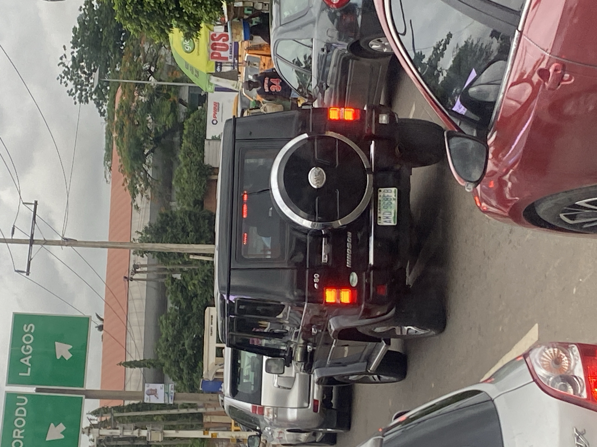 This image shows a traffic jam on a street in Ikeja, Nigeria. The foreground features the rear of a red car. Behind it is a black Innoson 4WD SUV with the license plate "AKD 968FB". To the left of the SUV is a silver Toyota SUV. In the background, traffic continues, and overhead signs indicate directions to "ORODU" and "LAGOS". Other elements visible include a street light, trees, buildings, and various other vehicles. The overall scene depicts a typical urban traffic situation.
