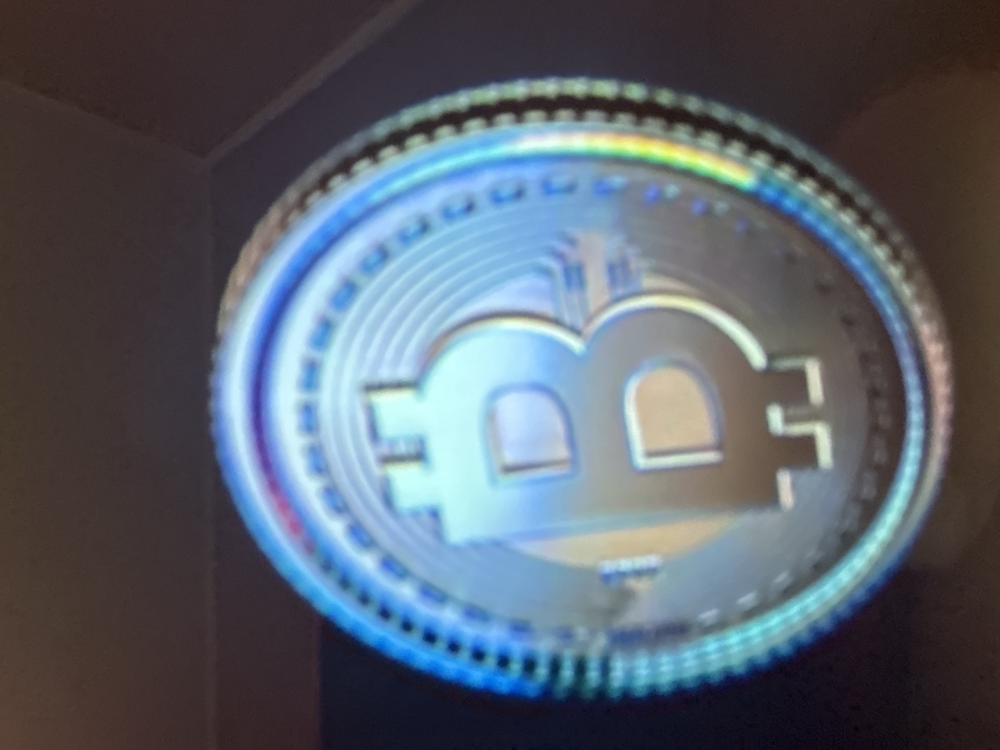 The image is a close-up, out-of-focus shot of a metallic Bitcoin coin. The coin is round with a distinct "B" symbol in the center, characteristic of Bitcoin. The surface of the coin appears to have a holographic or iridescent quality, reflecting light in blues, purples, and hints of yellow and green. The edge of the coin has a textured, ridged pattern. The background is dark and indistinct, making it impossible to discern any environmental details or a specific location. There is no information in the image to suggest a connection to Gelendzhik, Russia.