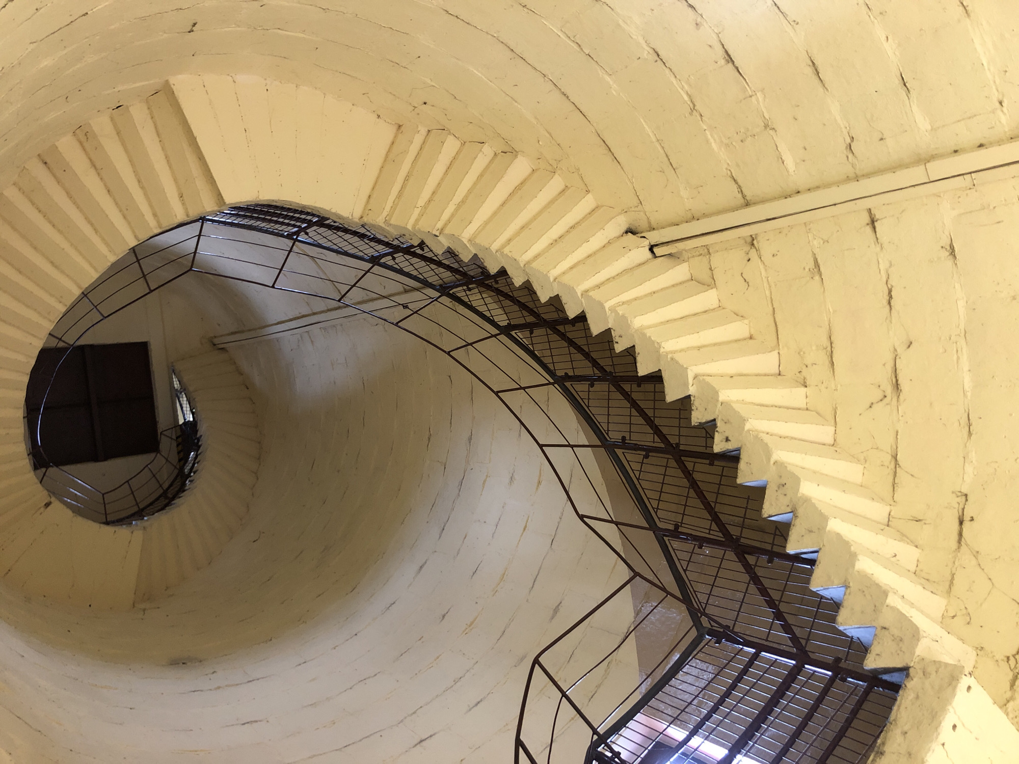 A low-angle, wide shot captures a beige, spiral staircase inside a cylindrical structure. The staircase is constructed from stone or concrete with a rough, textured surface and is illuminated by natural light filtering in from an unseen source, casting subtle shadows on the steps. A dark metal railing with a criss-cross pattern lines the outer edge of the staircase, curving upwards and inwards. The central column of the structure is visible in the background, forming a dark, circular opening at the top. The scene is devoid of people or any other discernible objects. The overall impression is one of austere, architectural grandeur.
