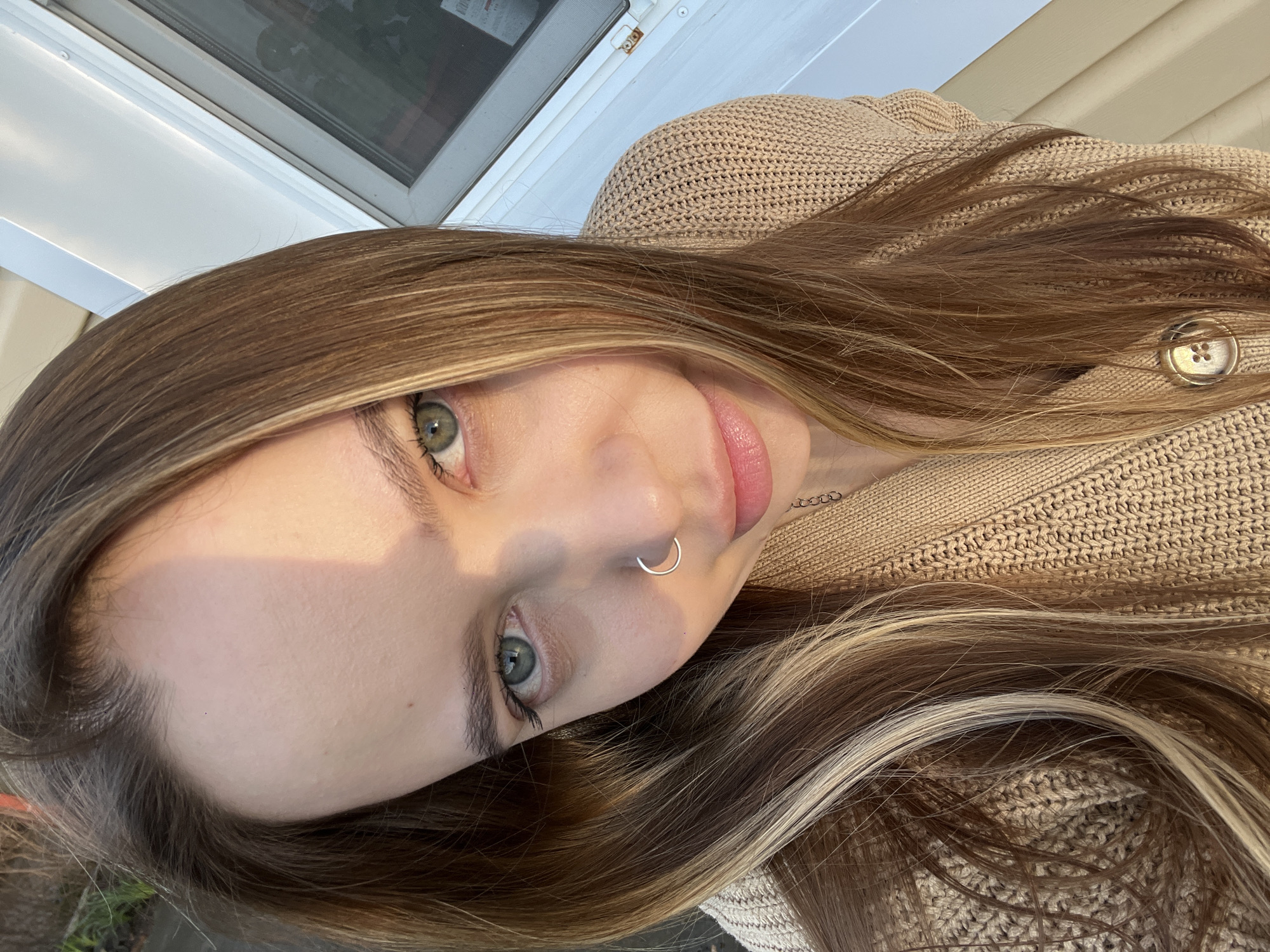 A close-up, rotated image shows a woman with long, brown hair with blonde highlights. She has fair skin, green eyes, and a silver nose ring. She is wearing a beige knit sweater with a button visible. The background includes a white window frame and a hint of green foliage. The location is not discernible as Novopokrovskaya, Russia, based solely on the visual information provided.