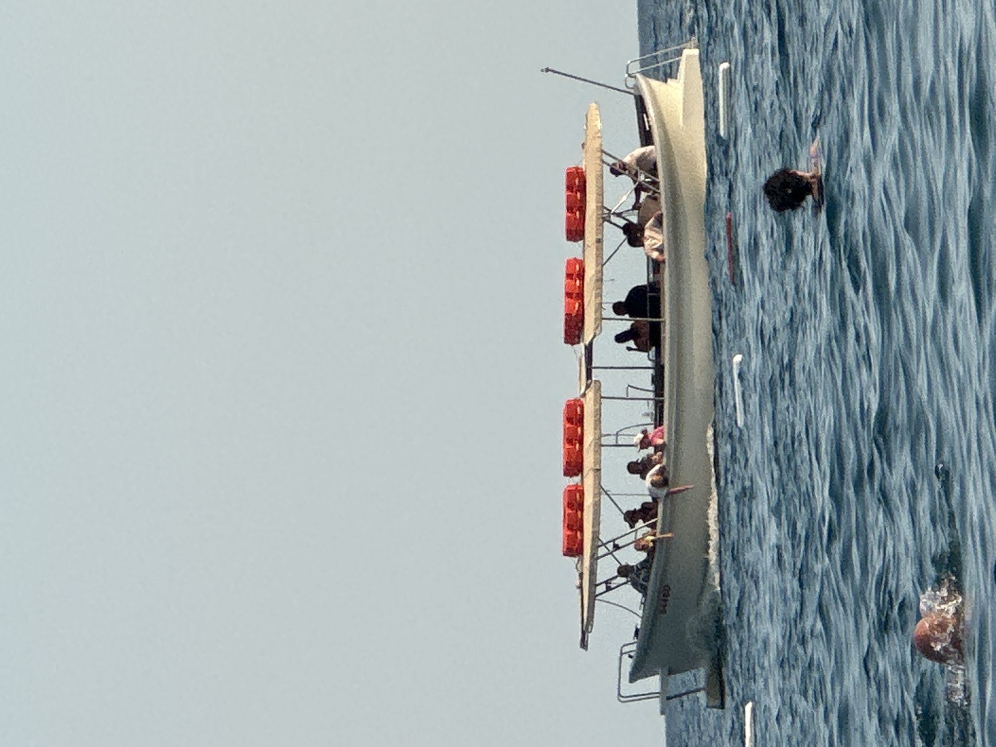 A light blue sky with a few wispy clouds is above a wide, off-white boat with a canopy. The boat, which appears to be a tour boat, has two red cylindrical life preservers attached to the top of the canopy. Several people are visible on the boat, including a person leaning over the side. The boat is on a blue ocean with gentle waves. In the foreground, a person with a bald head is swimming in the water, and another person with dark curly hair is also swimming. A white float is visible in the water near the boat. There are no visible landmarks in Boreti, Montenegro, in this image.