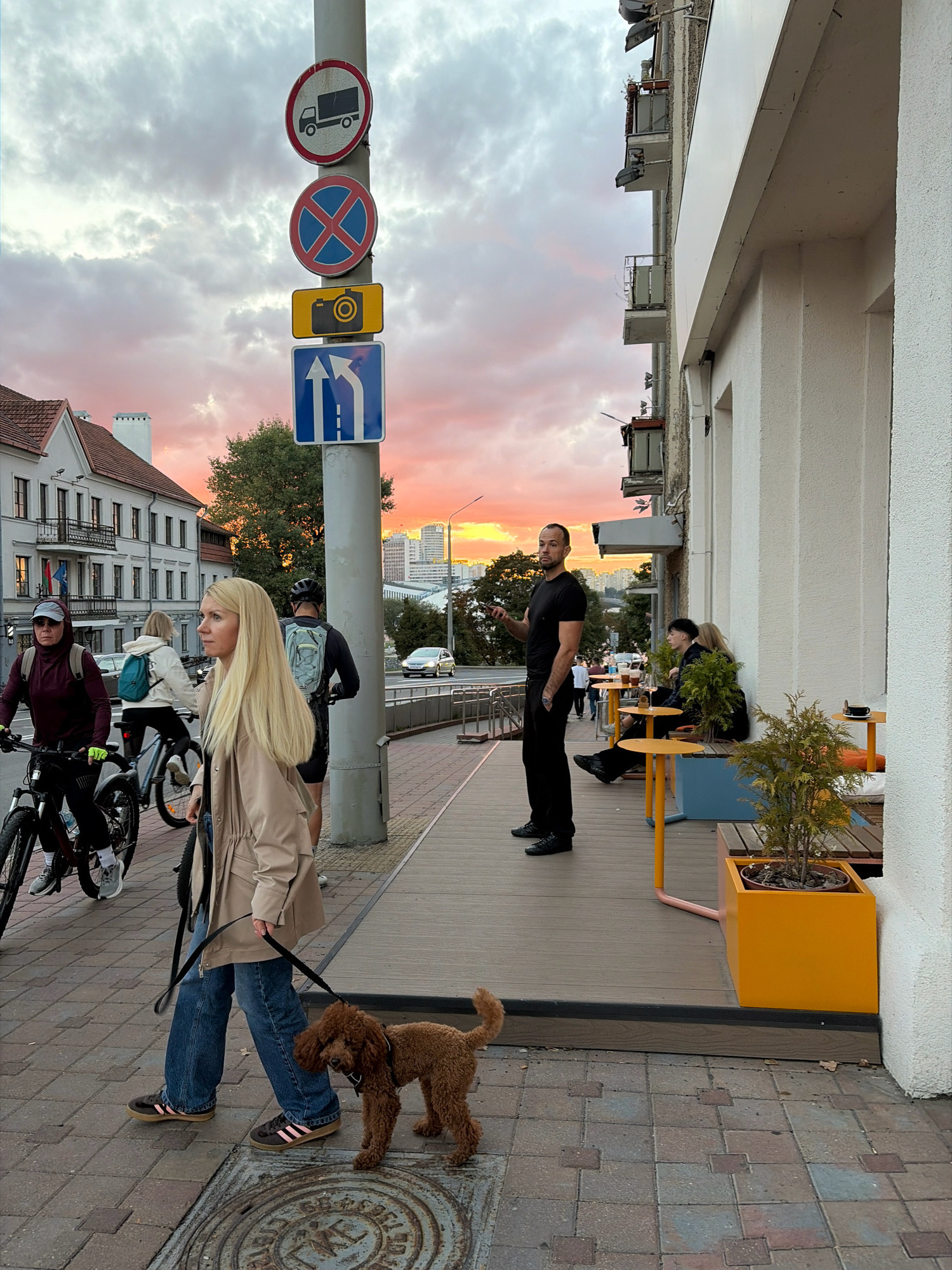In Minsk, Belarus, at dusk, a woman with long blonde hair, wearing a beige jacket and blue jeans, walks a small brown poodle on a leash. She is on a sidewalk paved with brick-like stones. Behind her, two individuals on bicycles pass by. A man in a black t-shirt and black pants stands on the sidewalk, holding a mobile phone. Further down the sidewalk, several people are seated at small yellow tables outside a building with white walls and balconies. The sky displays a colorful sunset with hues of pink and orange clouds. Traffic signs are visible on a pole near the sidewalk.
