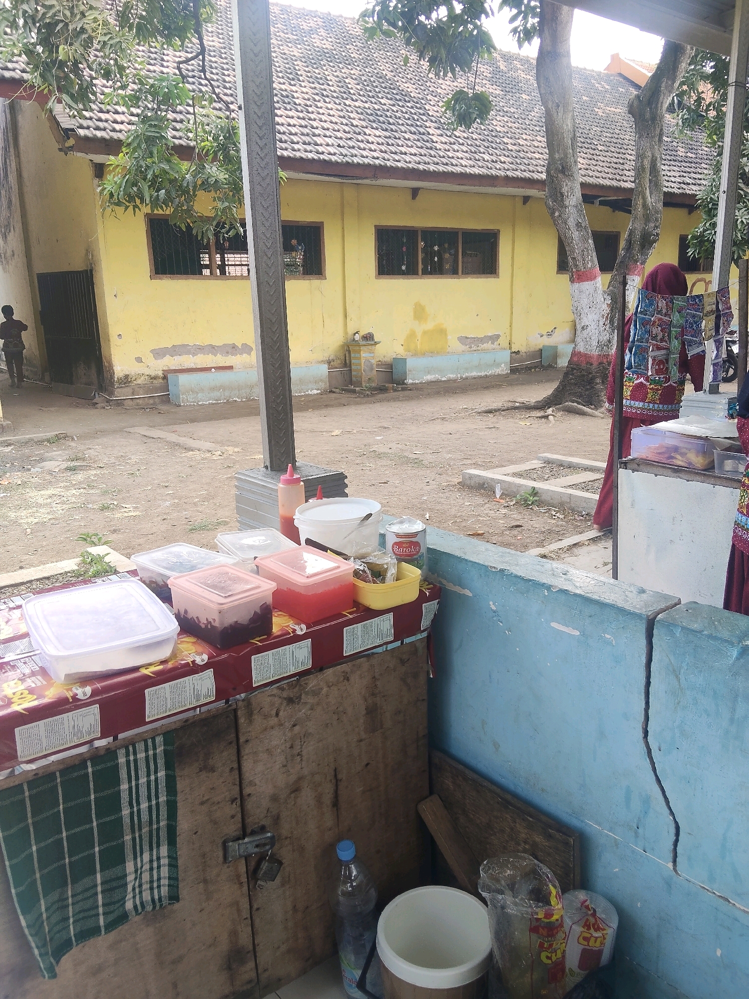 The image shows an outdoor scene in Payungrejo, Indonesia, near a yellow building with a tiled roof. In the foreground, there is a food stall with several plastic containers filled with food items. A red and white checkered cloth hangs on a wooden cabinet. There are also bottles of water and other containers on the ground. A person wearing a headscarf and a colorful outfit stands behind the stall, facing away from the camera. The ground is covered in dirt and gravel. In the background, trees and the yellow building are visible. The overall scene depicts a casual, possibly rural, setting with a focus on a food vending operation.