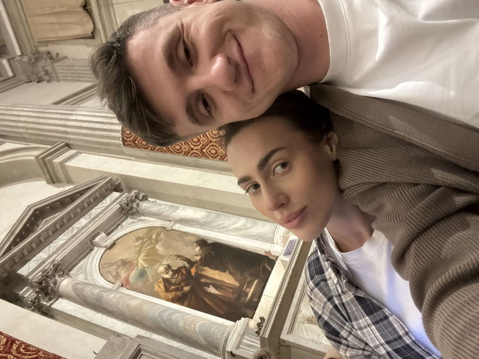 A couple is captured in a close-up, selfie-style photograph inside the Chiesa di San Zaccaria in Venice, Italy. The man, positioned on the right, has short dark hair and wears a white t-shirt, looking at the camera with a slight smile. The woman, on the left, rests her head on the man's right shoulder, gazing neutrally at the camera. She has dark hair and wears a white t-shirt under a plaid shirt and a brown cardigan. The background features a large, ornate baroque altar structure with fluted marble columns, Corinthian capitals, an entablature, and a pediment. Within this frame is an oil painting, "The Calling of Saint Matthew" by Jacopo Palma il Giovane, depicting an elderly robed figure gesturing upwards towards celestial beings, with another figure kneeling below. The surrounding church walls are adorned with tall, fluted columns and patterned red and gold fabric.