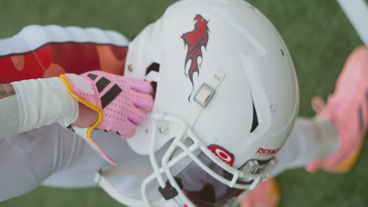 An extreme close-up shot shows a person's hand in a pink and yellow Adidas glove, holding a white football helmet with a red dragon decal on the side. The helmet has a white face mask. The person's arm is wrapped in white tape. They are wearing white pants and pink cleats. The background is blurred green grass. The image is tightly cropped, focusing on the equipment and hand.
