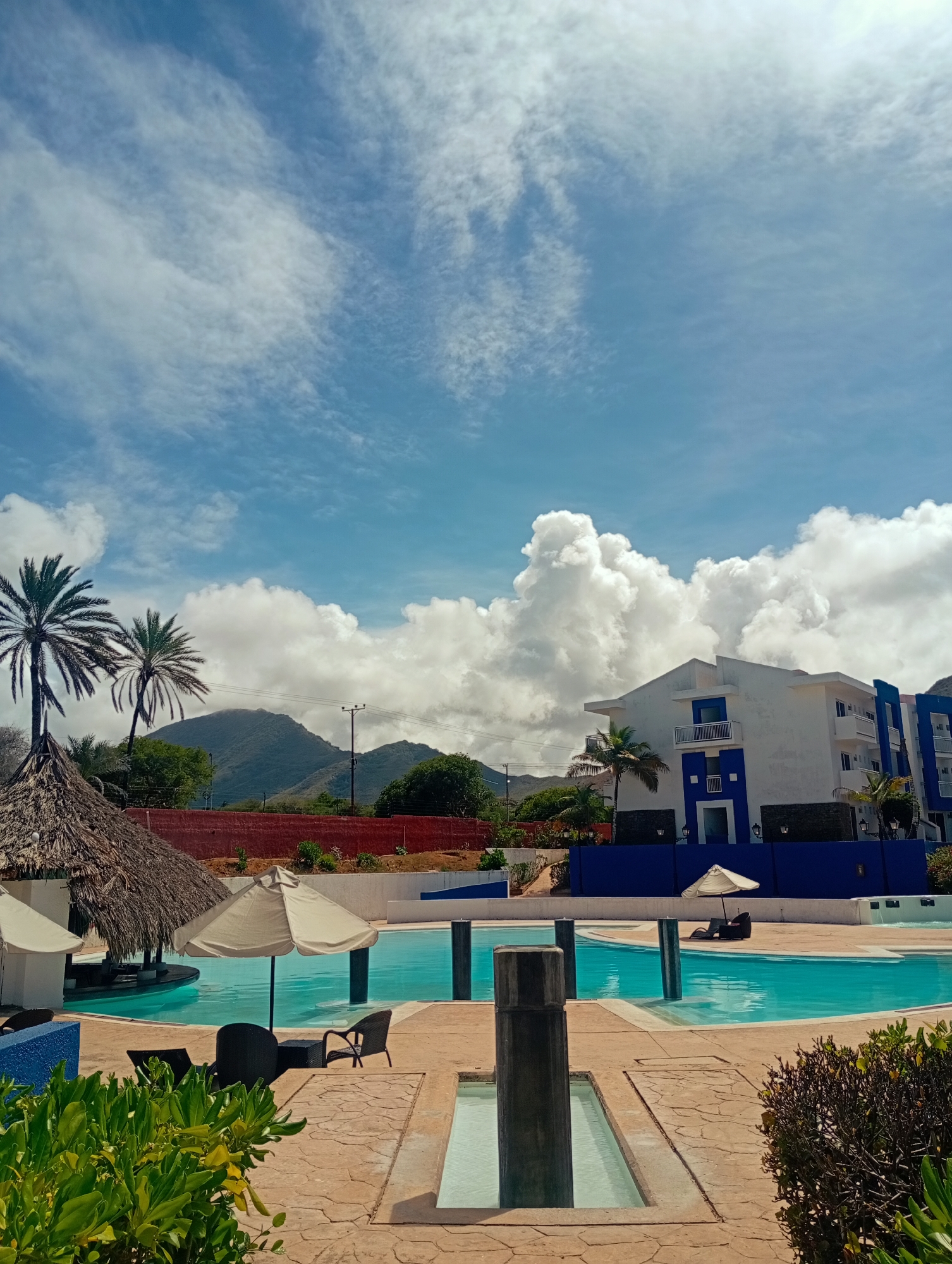 The image depicts a resort-style pool area in Playa El Agua, Venezuela, set against a backdrop of mountains and a partly cloudy sky. The pool, filled with turquoise water, is surrounded by a textured tan patio. In the foreground, there is a decorative shallow water feature lined with stone and greenery. Several gray columns rise from the pool's surface.
To the left, a thatched-roof bar stands next to two palm trees. Beige patio umbrellas and dark wicker furniture provide seating areas. A building with white walls and blue accents is visible behind the pool. Further in the background, a rusty red wall sits in front of tree covered mountains. No people are present in the scene.