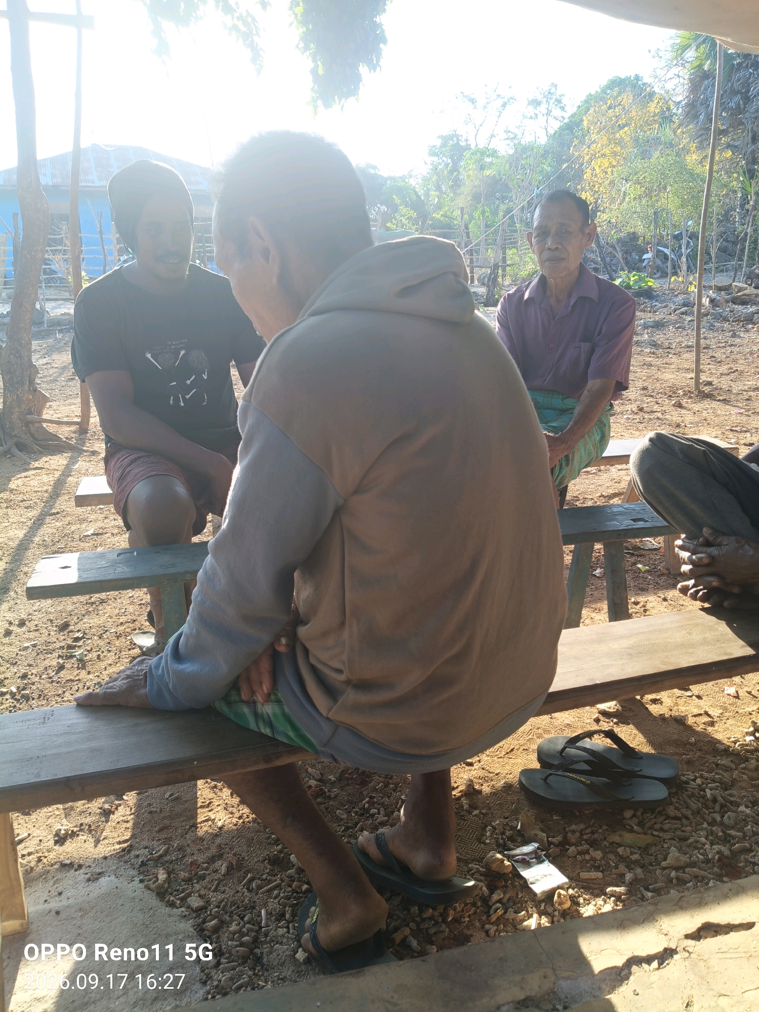 The image depicts an outdoor scene, likely in Otan, Indonesia, where three men are seated on wooden benches. The lighting suggests it is daytime, with strong sunlight casting shadows.
In the foreground, a man is seated on a bench, with his back mostly to the camera. He is wearing a light brown hooded sweatshirt and shorts. His legs are bare, and he is wearing sandals. His right hand rests on the bench.
To his left, another man is seated on a separate bench, facing the first man. He is wearing a dark t-shirt and patterned shorts. He appears to be in conversation with the man in the brown sweatshirt.
In the background, a third man is seated on a bench facing the camera. He is wearing a purple collared shirt and a green and white checkered sarong. He looks towards the camera with a neutral expression.
The environment appears to be somewhat rustic. The ground is covered with sand and small stones. Behind the men, there are trees and vegetation, and a simple structure that could be a house or shelter. The overall impression is of a casual gathering in a rural or semi-rural setting.