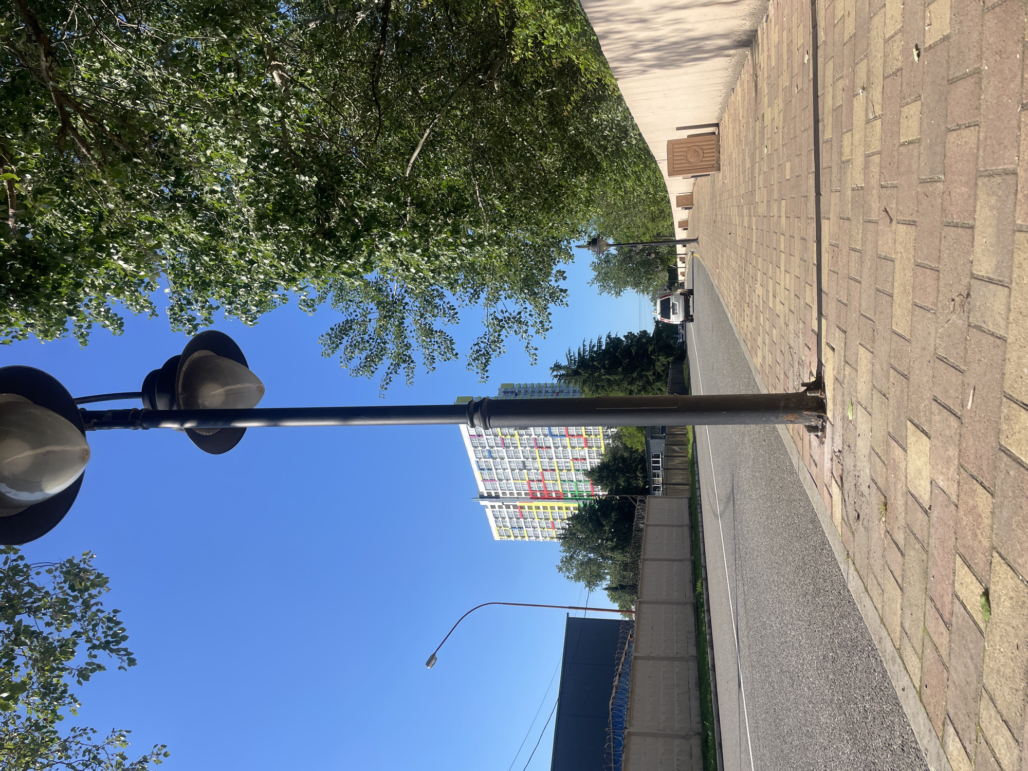 A bright, sunny street scene unfolds under a clear blue sky, likely in Sochi, Russia. In the foreground, a prominent black streetlamp with two white, bowl-shaped lights stands on a paved sidewalk. The sidewalk is constructed from rectangular bricks in shades of light brown and reddish-brown, extending into the distance alongside an asphalt road. Lush green tree canopies line the left side of the street, casting shadows.
Further down the road, on the right, a low concrete fence topped with barbed wire runs parallel to the street. Behind this fence, a large, multi-story building stands out with a distinctive facade. It features a predominantly white exterior adorned with a vibrant, grid-like pattern of colorful panels or window frames in red, yellow, green, and blue, suggesting a modern residential or commercial structure.
A smaller, arching streetlamp is visible above the road in the mid-distance. A white van is partially visible on the road further back. Several square, brown metal covers, possibly for utility access, are embedded in the sidewalk. Each cover displays a circular emblem with a discernible design, though any accompanying text is too faint and blurry to be deciphered. The clear lighting and visible shadows indicate it is daytime, likely mid-morning or afternoon, with pleasant, clear weather. No specific human activity is depicted.