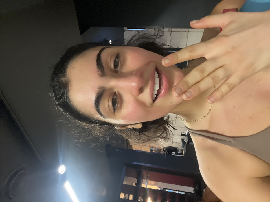 A selfie of a young woman with dark hair pulled back. She is smiling and facing the camera. Her skin appears dewy. She is wearing a simple necklace. One hand is raised near her face. The background is dark, suggesting an indoor setting with some architectural elements visible.
