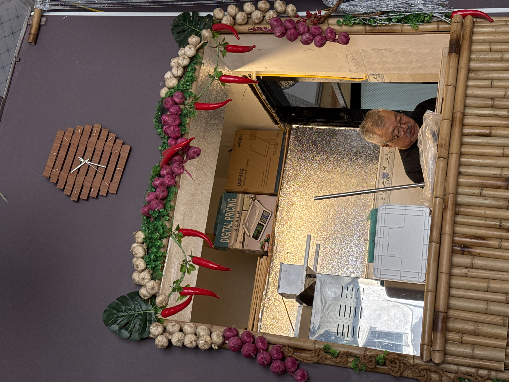 The image shows a man in a black shirt and glasses with a gray mustache and beard, wearing round eyeglasses, looking down and reading at a counter made of bamboo. He is in what appears to be a food stall or small eatery. The stall is decorated with garlands of artificial garlic, red chili peppers, and purple onions. Behind him, there is a reflective surface, and a box labeled "DIGITAL PRICING SCALE" and "IAP302 Touch Screen POS System" is visible. The overall impression is of a rustic, possibly Vietnamese, establishment. The location is identified as a food stall in Nha Trang, Vietnam.