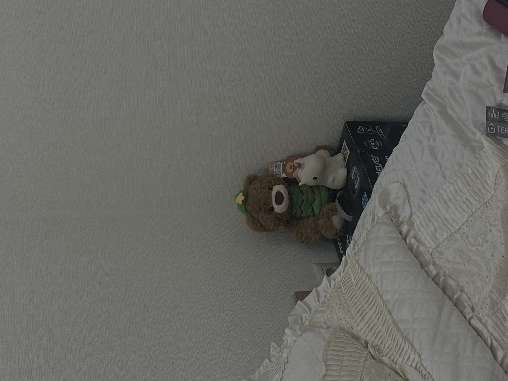 The image shows a bedroom scene, likely indoors, with a light gray wall on the left side taking up a significant portion of the frame. Towards the right of the frame, a bed with white bedding featuring intricate lace and pleated details is visible. On the bed, there is a black box and a brown teddy bear wearing a green scarf with yellow clover or flower. On top of the bear is a small, white, possibly plastic, animal figurine. The top right corner features a red item and packaging, indicating the presence of other objects on the bed. The scene is cluttered and domestic, suggesting a lived-in space. There is no information available to suggest this is Yerevan, Armenia.