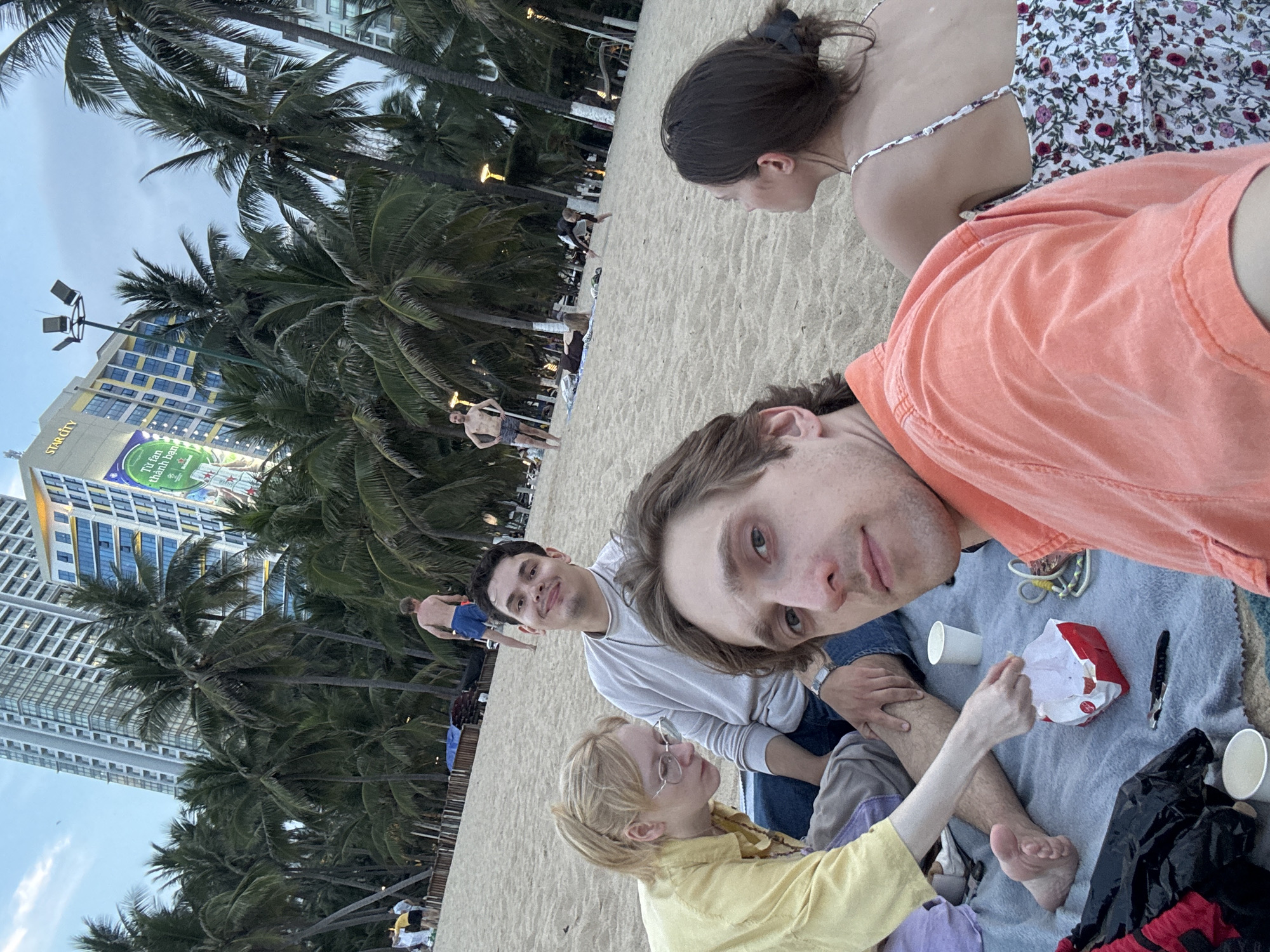 A group of people are sitting on a beach blanket on the sand, with palm trees and modern buildings in the background. The image appears to be taken in Nha Trang Ward, Vietnam, as suggested by the architecture and tropical setting.
In the foreground, a man in an orange shirt is looking at the camera, with his face filling a significant portion of the frame. To his left, a woman with blonde hair and glasses, wearing a yellow shirt, is looking down, reaching towards a red and white food container. Next to her, a man in a light gray shirt is looking at the camera and smiling. Further back, a woman with dark hair in a floral dress is facing away from the camera.
In the mid-ground, several other people are visible on the beach, some walking and others relaxing. The background features a dense line of palm trees with buildings of varying heights behind them, including a tall, modern skyscraper with a prominent "STAR CITY" sign. The sky is overcast.
Objects visible on the blanket include two white paper cups, the red and white food container, a knife, and a rope. The overall impression is a casual gathering on a beach, likely in the late afternoon or early evening given the lighting.