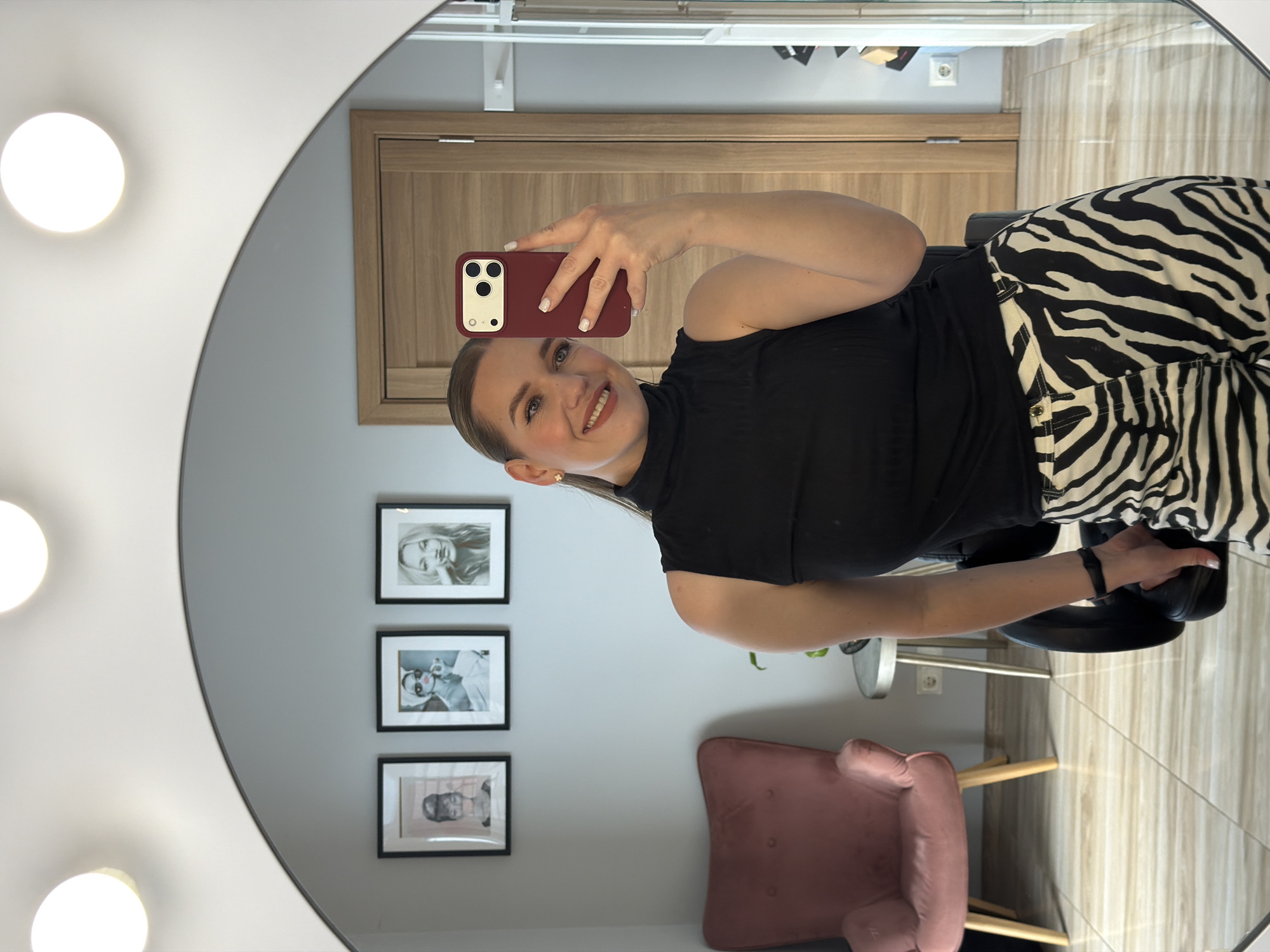In Riga, Latvia, a woman takes a selfie in a well-lit room, reflected in a large, arched mirror. She is wearing a black sleeveless turtleneck and black and white zebra-striped pants. Her hair is pulled back into a ponytail, and she is smiling, holding a red smartphone in her right hand. The mirror reflects a dusty pink armchair, three framed black and white photographs on a light gray wall, a wooden door, and a tall, narrow cabinet. The floor is tiled with a wood-grain pattern. The scene appears to be a dressing room or a boudoir.