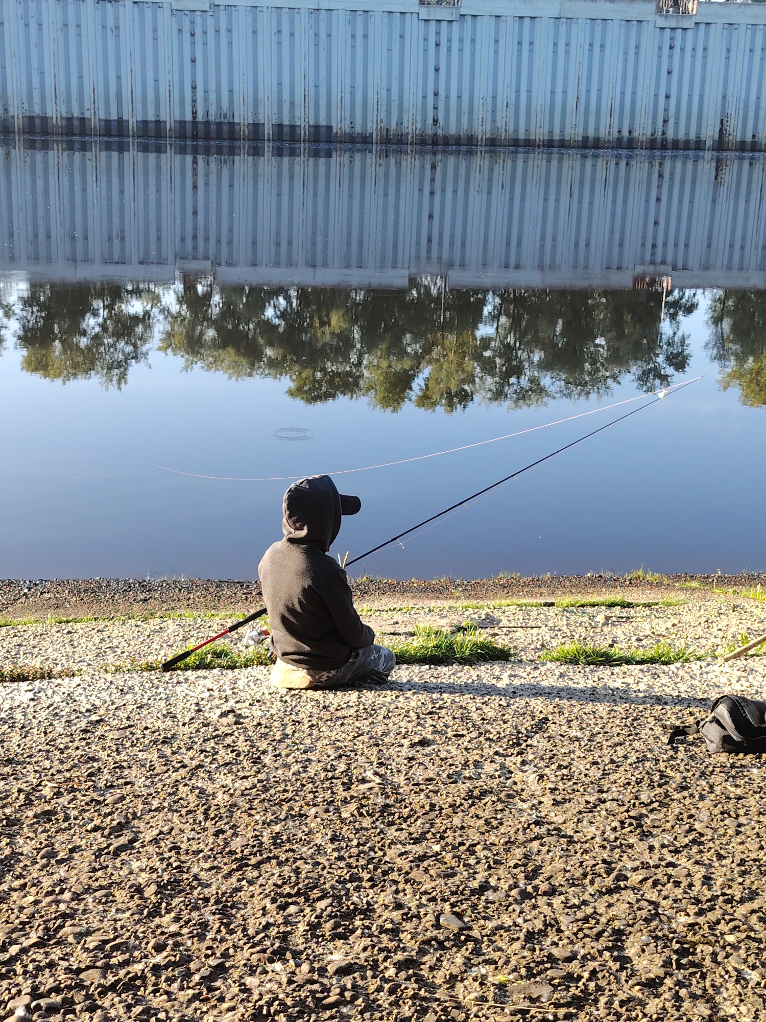 A person wearing a dark hooded sweatshirt and a baseball cap is sitting on the gravel bank of a body of water, facing away from the camera. The person is holding a fishing rod that extends into the water. The water reflects a tall, grey, ribbed structure that runs along the opposite bank. Trees are also reflected in the water. The ground in the foreground is covered in small stones and sparse green grass. A black backpack is visible to the right of the person. The scene appears to be in a park or recreational area along the Kama River in Chaikovsky, Russia.