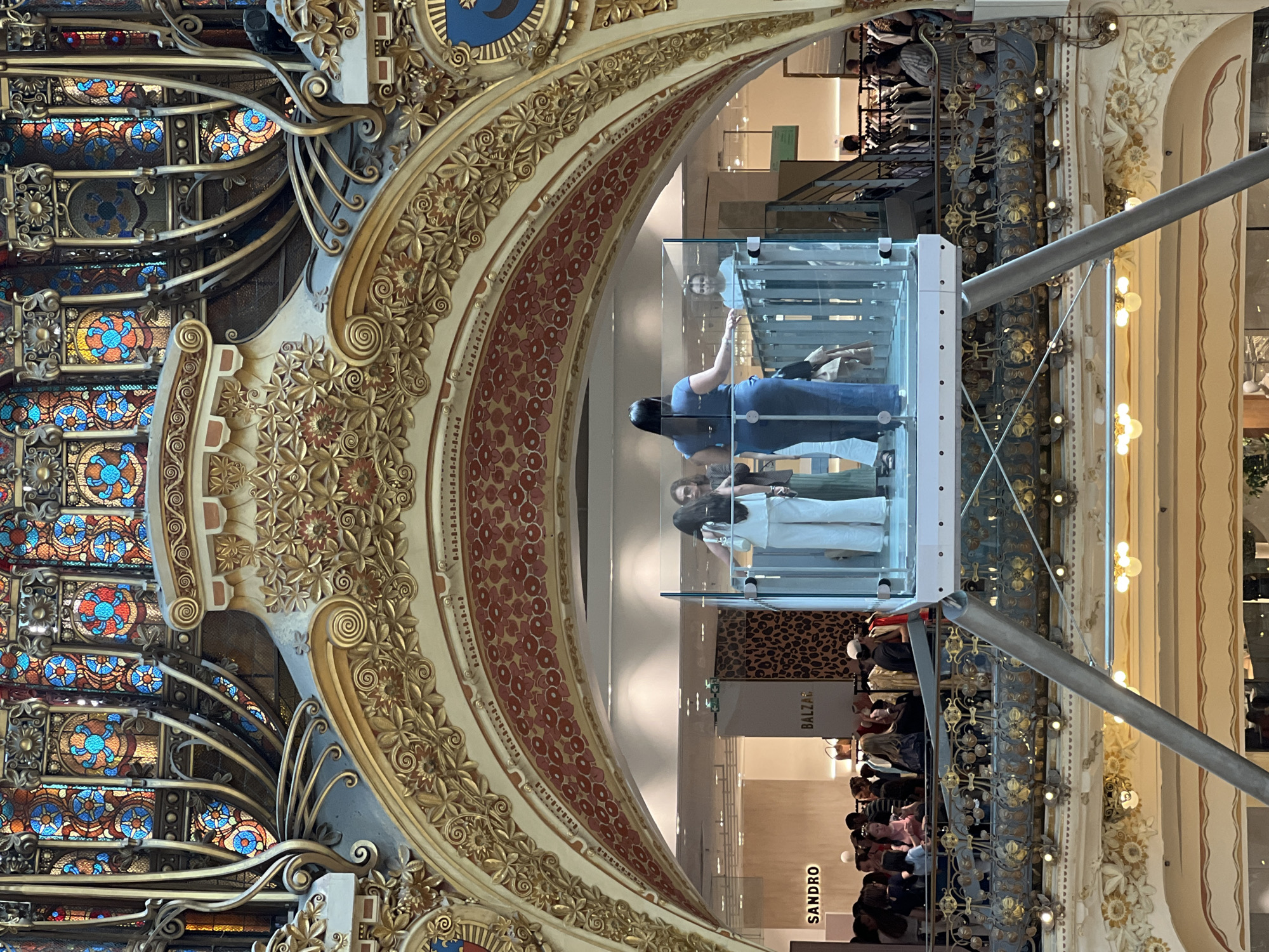 The image shows the interior of the Galeries Lafayette Haussmann department store in Paris, France. The focal point is an ornate, art nouveau-style glass elevator with people inside, viewed from below through an archway decorated with gold ornamentation and stained glass.
In the foreground, a metal beam from the elevator structure extends diagonally upwards. The elevator itself is enclosed by glass panels, revealing three individuals. The person closest to the viewer, on the left, is a woman wearing a white top and white pants. Next to her is another woman in a dark top and dark pants. On the right side of the elevator is a woman in a blue dress.
Through the glass elevator and the archway, the department store's interior is visible. There are retail displays with clothing racks and mannequins. Signs for "SANDRO" and "BALZAI" are discernible. Other shoppers are seen browsing the merchandise on the floor below. The architecture of the store is elaborate, featuring decorative gold trim, floral motifs, and stained-glass windows at the top of the frame. The lighting suggests an indoor environment. The overall impression is one of grandeur and commercial activity within a historically significant building.