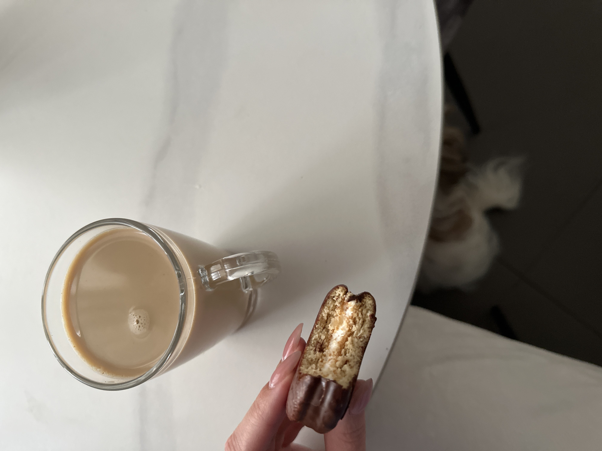 A top-down shot depicts a clear glass mug containing a light brown beverage, likely coffee or latte, resting on a white, marble-patterned surface. Beside the mug, a hand with long, manicured nails holds a partially eaten, dark chocolate-covered snack cake with a visible cream-filled interior. The background is blurred, but what can be seen suggests a room interior with a white surface and dark wall. The location cannot be determined from the information available in the image.