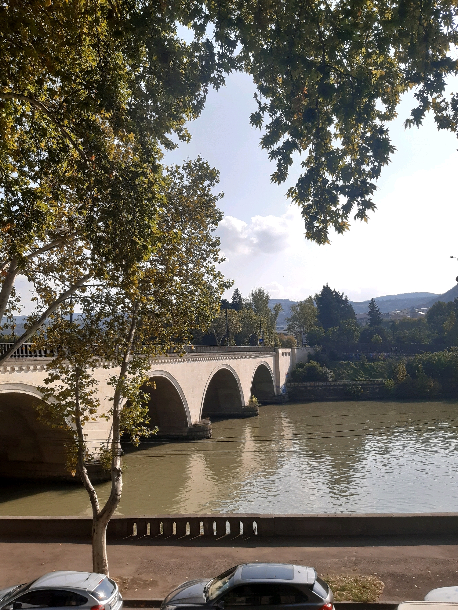 The image depicts a view of the Kura River in Tbilisi, Georgia, with a stone bridge with multiple arches spanning across it. The bridge is light-colored and features decorative elements along its upper edge. The river's surface is a muted, greenish-brown, reflecting the overcast sky.
In the foreground, a paved embankment runs along the river, lined with a low, decorative stone wall. A slender tree with light-colored bark and sparse green and yellow leaves stands near the embankment. To the left and right of this tree, portions of two parked cars are visible on a street adjacent to the embankment. The car on the left is a silver hatchback, and the car on the right is a dark gray hatchback.
The background reveals a lush, green hillside covered with various trees. In the distance, the silhouette of hills and some buildings can be seen, suggesting the urban landscape of Tbilisi. The sky is bright with some scattered clouds. The overall scene suggests a tranquil, daytime setting in an urban environment, likely during the autumn season given the foliage colors.