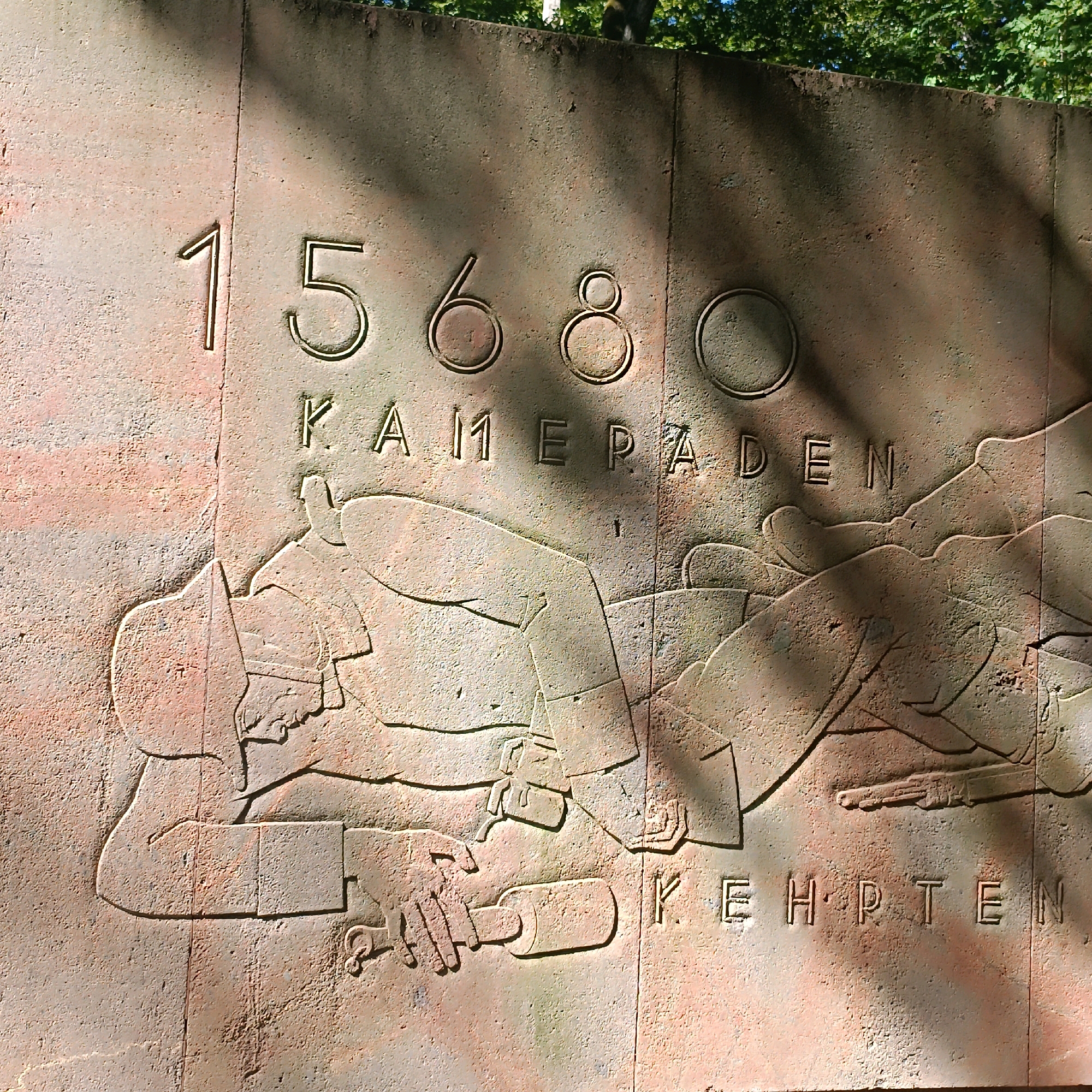 The image depicts a bas-relief sculpture carved into a textured, reddish-brown stone wall. The sculpture is divided into two main sections by a vertical line down the center of the image.
The upper section prominently displays the numbers "15680" in large, raised capital letters and Arabic numerals, with each digit enclosed in a circular outline except for the '1' and '5'. Below this, the word "KAMERADEN" is carved in capital letters, likely representing "Comrades" in German.
The lower section of the sculpture features figures in relief, appearing to depict a scene of hardship or struggle. On the left, a figure is shown lying down, reaching out with one hand towards a bottle or flask, which is being held by another figure. This second figure is also lying down and appears to be offering the object. The figures are depicted wearing garments that suggest a historical or military context, with details like collars and headwear visible. A rifle is also present, lying across the body of the figure on the right.
To the right of the central vertical line, the word "KEHRP.TEN" is carved in capital letters. The overall style of the sculpture is somewhat stylized and monumental. The wall itself appears to be part of an outdoor structure, with dappled sunlight and shadows from foliage falling on its surface, indicating an exterior setting, possibly a memorial.
This sculpture is located at the "Rathaus" (City Hall) in Wiesbaden, Germany, and is part of the "Friedhof der Namenslosen" (Cemetery of the Nameless). It commemorates victims of the Thirty Years' War.