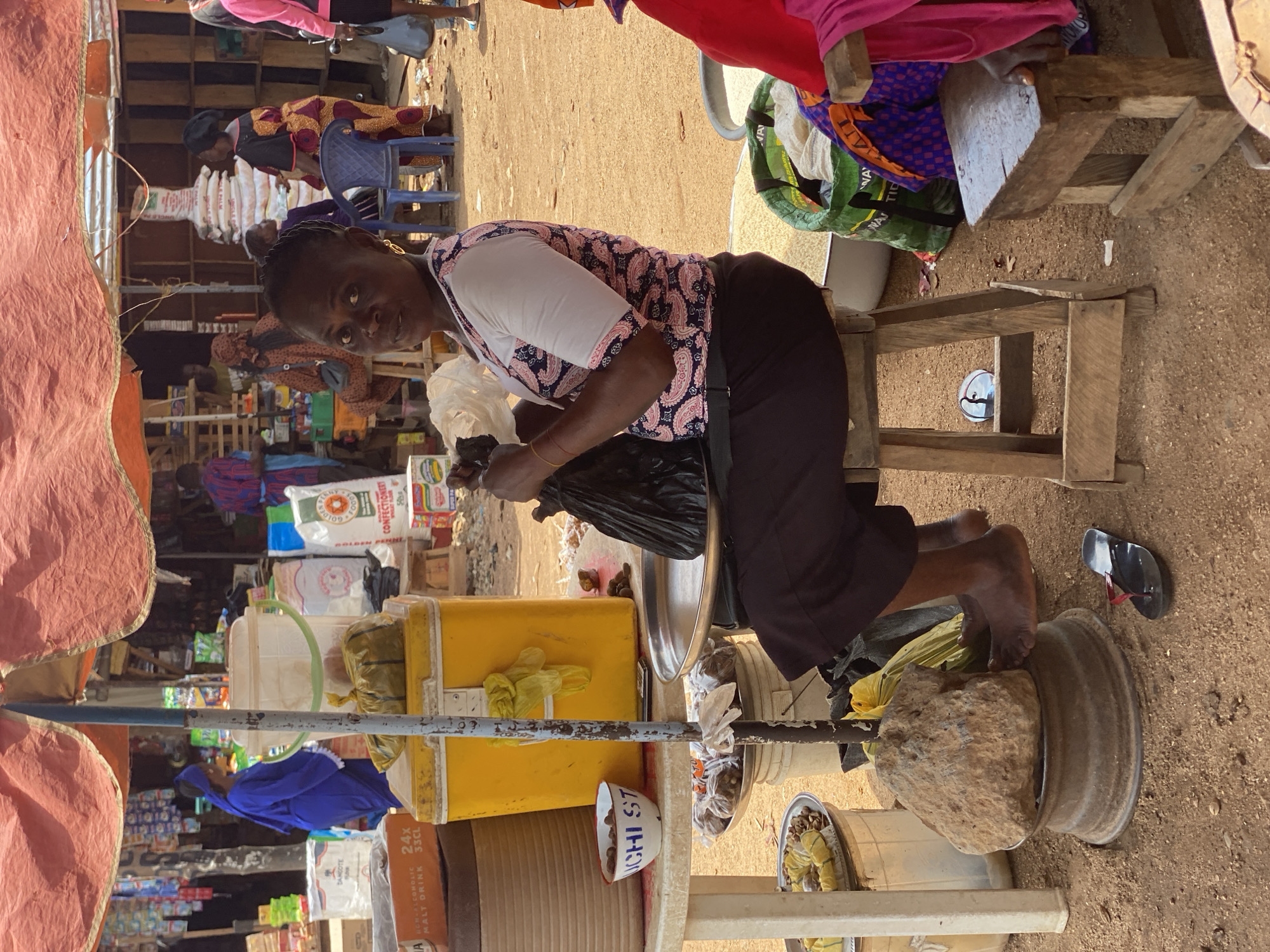 This image captures a vibrant market scene in Kafanchan B, Nigeria, during bright daylight. The main subject is a woman, clad in a patterned white and black blouse and dark trousers, kneeling barefoot on a low wooden stool. She looks directly at the camera with a neutral expression while appearing to handle goods, possibly weighing items into a black plastic bag using a makeshift scale comprised of a large stone balancing on an inverted metal rim on the ground.
The setting is an outdoor market with unpaved, sandy ground and various makeshift stalls under fabric awnings visible on the left. In the background, several other individuals are present, some sitting or standing amidst numerous market items. The scene is bustling with merchandise, including large white sacks labeled "GOLDEN PENNY FLOUR CONFECTIONERY," a brown box for "DAMCOTE A NON-ALCOHOLIC MALT DRINK," and other packaged goods stacked on shelves or displayed in yellow and clear plastic containers. A white bowl with "ICHI ST" written on it is also visible. The woman's black flip-flops are placed beside her on the ground. The overall impression is one of a busy, informal commercial environment.