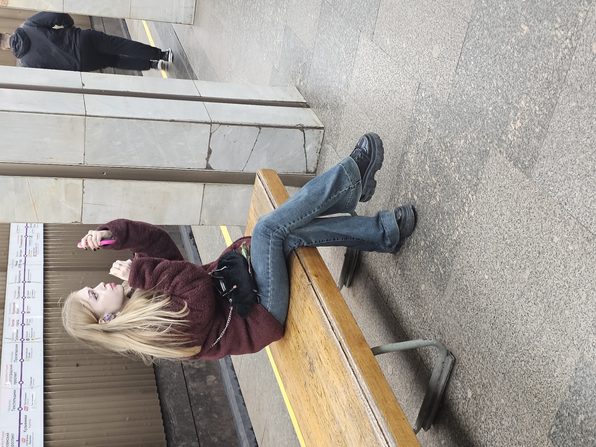 The image captures a relaxed moment within a Moscow Metro station. A young woman, the primary subject, is seen lying sideways on a wooden bench, engaged with her pink-cased phone. She has long blonde hair, wears a dark red fuzzy jacket, blue jeans, and distinctive black chunky-soled shoes. Several rings adorn her fingers, and her nails are painted dark. A small black bag or pouch with a chain rests beside her on the bench.
In the background, another individual, a man in a dark jacket and pants, is visible standing or walking on the platform. The station architecture features large, light-colored stone pillars and a speckled grey and light beige tiled floor, with a yellow safety line running along the platform edge.
An information sign, likely a subway map or station list, is mounted on the wall behind the woman. Visible text on this sign, written in Cyrillic, lists several Moscow Metro stations. These include "Кузьминки" (Kuzminki), "Таганская" (Taganskaya), "Пролетарская" (Proletarskaya), "Волгоградский проспект" (Volgogradsky Prospekt), "Текстильщики" (Tekstilshchiki), "Рязанский проспект" (Ryazansky Prospekt), "Китай-город" (Kitay-gorod), "Пушкинская" (Pushkinskaya), "Баррикадная" (Barrikadnaya), "1905 года" (1905 Goda), "Беговая" (Begovaya), and "Полежаевская" (Polezhaevskaya). The setting indicates an indoor public space, likely during the day or under bright artificial lighting. The overall mood is casual, showing the woman waiting comfortably. The photo is rotated 90 degrees clockwise.
