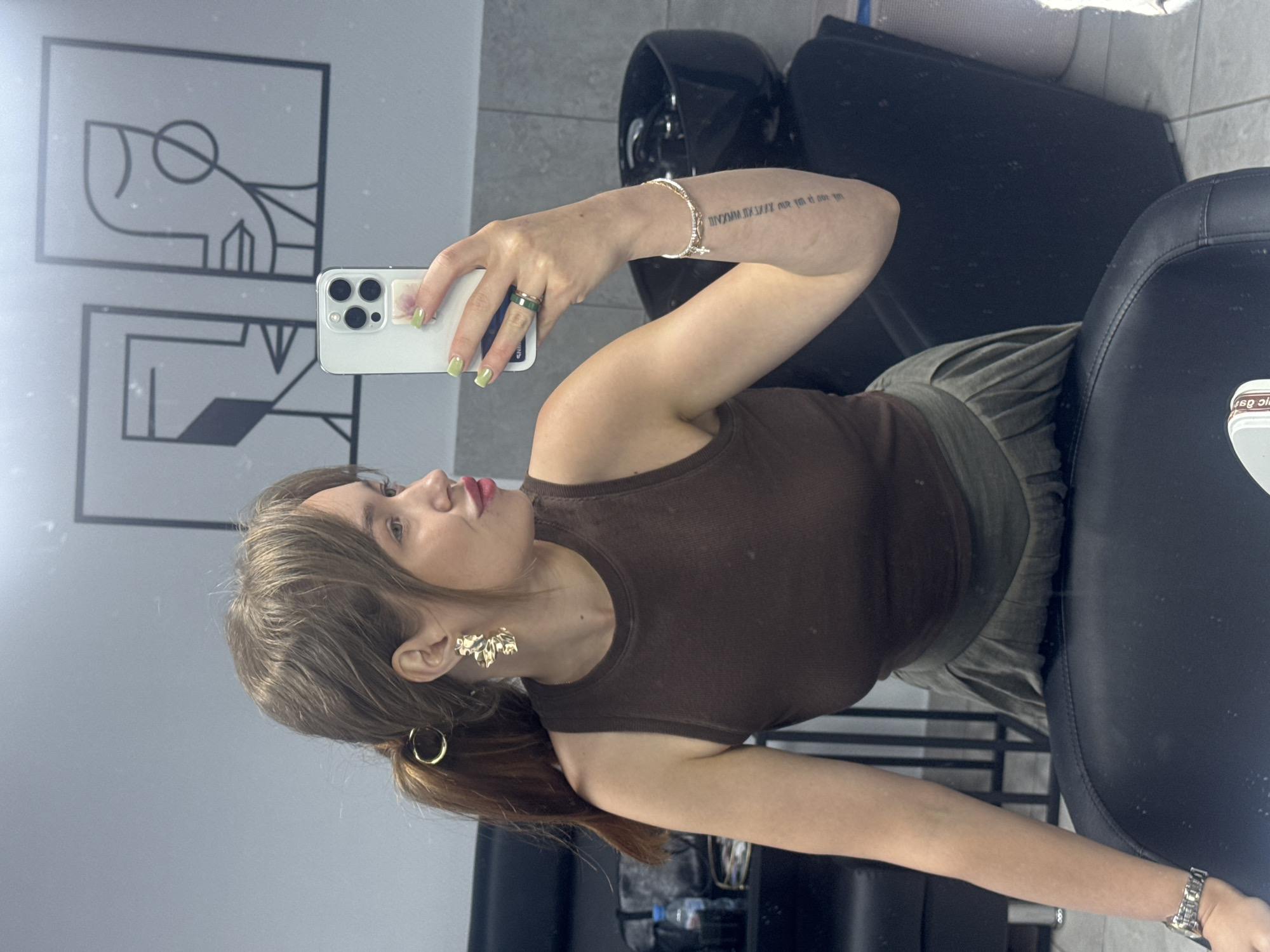 A woman is taking a selfie in a salon in Chaikovsky, Russia. She has light brown hair pulled back in a ponytail with a gold hair accessory. She is wearing large gold earrings, a brown tank top, and a green pleated skirt. She is also wearing a green ring and a bracelet on her right hand. A tattoo is visible on her left forearm. The background shows a tiled wall with two abstract framed pictures, a black salon chair, and a sink.
