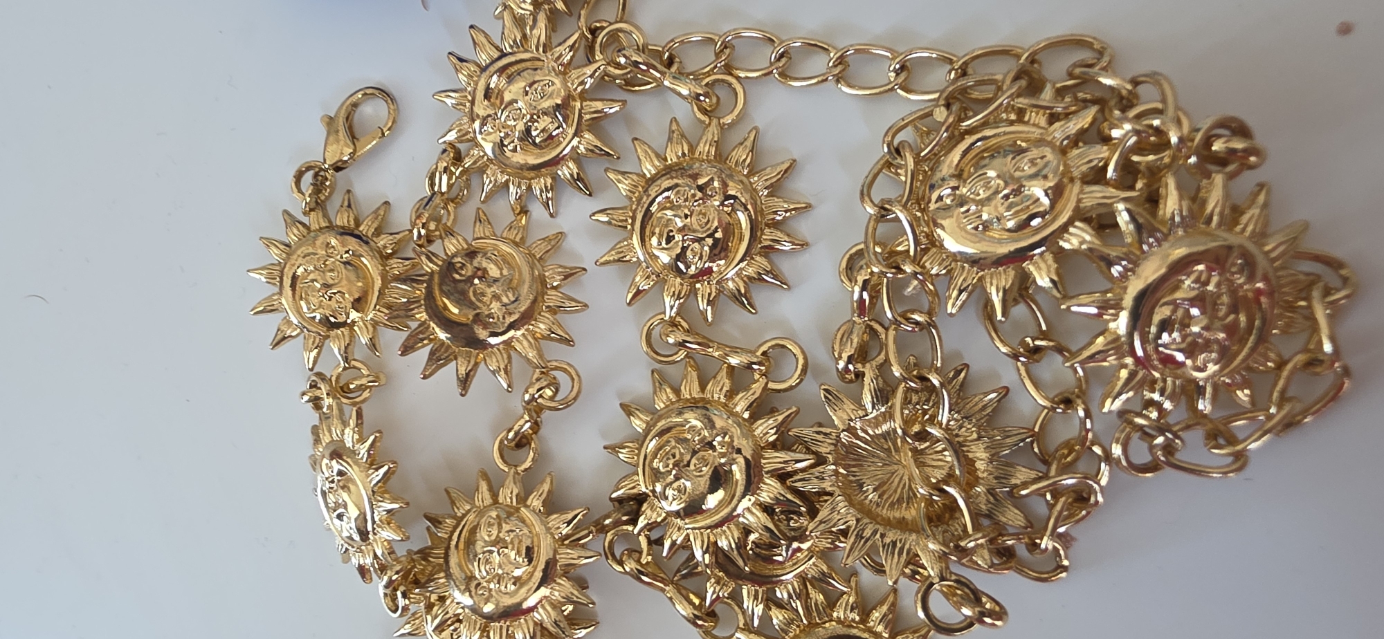 The image shows a close-up, eye-level shot of a gold-tone chain belt adorned with multiple sun-shaped charms featuring faces. The belt is laid out on a plain, white surface. The sun charms have intricate details with radiating rays and expressive faces with eyes, noses, and mouths. The chain links are oval-shaped. The overall impression is of a piece of costume jewelry. There is no discernible urban or natural environment, and therefore no location in Sumgayit, Azerbaijan can be inferred.
