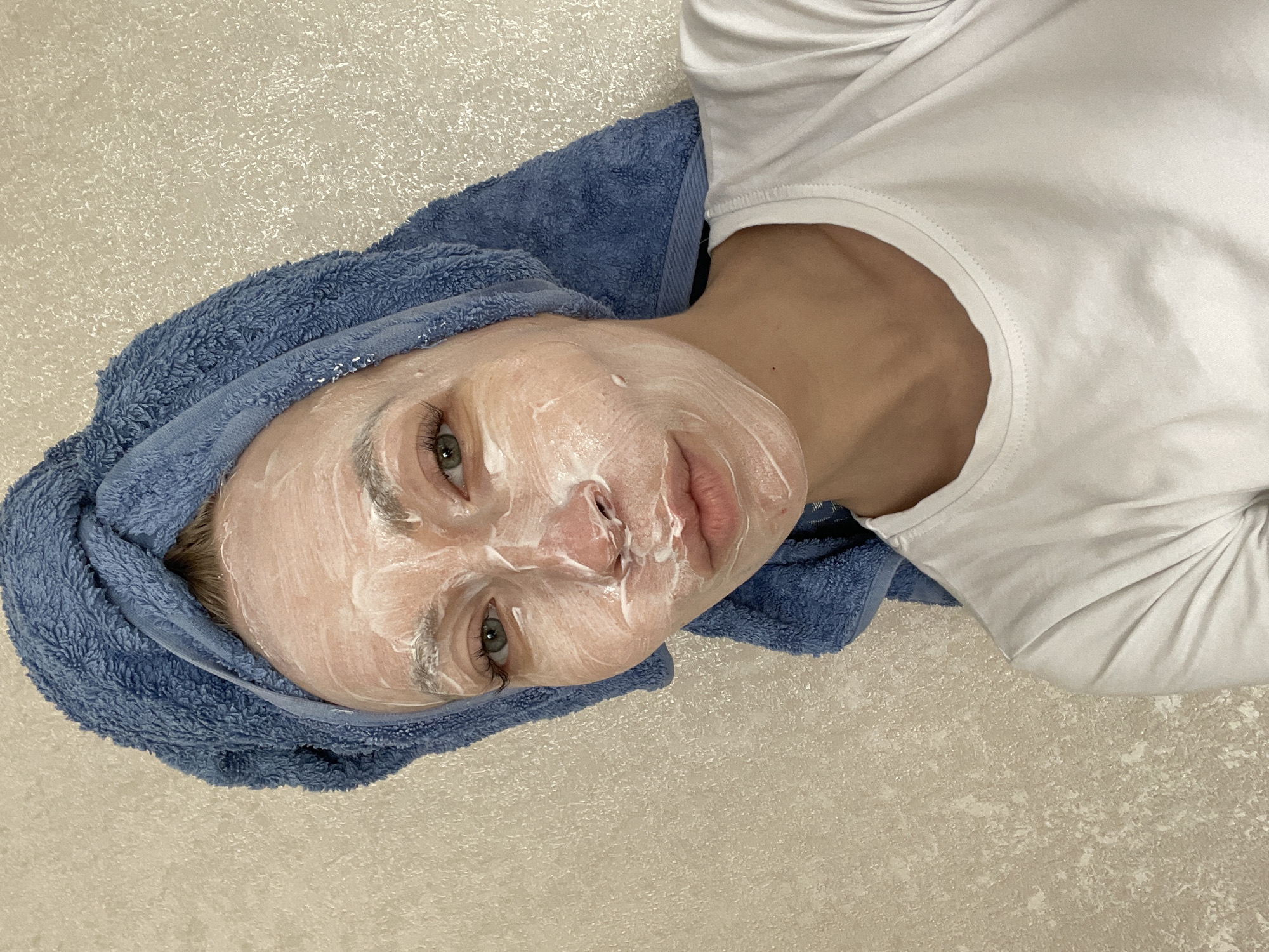 The image shows a close-up, diagonal view of a person engaged in a skincare routine, likely indoors. The subject, who appears to be an adult with fair skin and light-colored eyes, has their entire face covered in a thick, white creamy substance, resembling a facial mask or cleanser. A blue terry cloth towel is neatly wrapped around their head, holding their hair back. They are wearing a plain white short-sleeved t-shirt, visible on the right side of the frame.
The person is lying down, with their head positioned towards the upper left and their body extending diagonally towards the lower right. Their gaze is directed towards the camera with a neutral expression. The background is a uniform light beige or off-white textured surface, possibly wallpaper or a tiled floor, suggesting a clean and simple setting. There are no specific cues to indicate the time of day or weather. No visible text is present in the image. The scene captures a moment of personal care and relaxation.