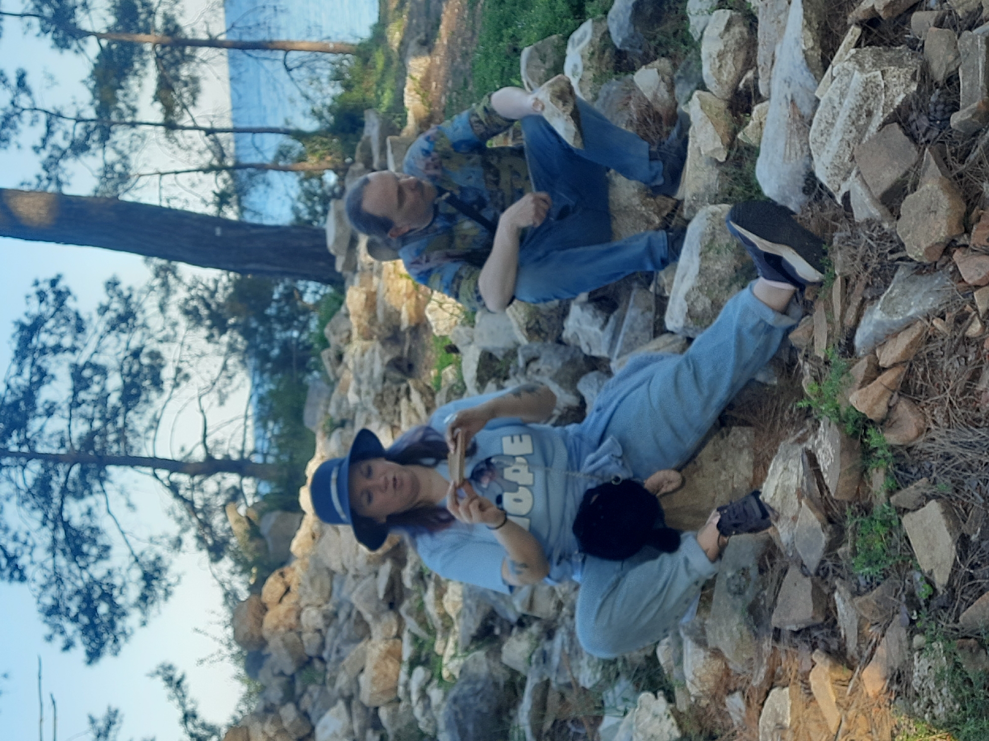 The image, rotated 90 degrees clockwise, portrays two individuals relaxing outdoors on a rocky incline by a body of water, set in Kemer, Turkey.
In the foreground, a woman is seen reclining on her side amidst light-colored rocks, sparse dry grass, and some green foliage. She is dressed in a light blue t-shirt with the letters "OPE" clearly visible, suggesting the word "HOPE." She also wears light blue pants, a dark blue hat with a light band, and dark athletic shoes. Tattoos are visible on her left arm as she holds a small, rectangular object, possibly a phone, with both hands. A small black bag or object rests near her.
Behind her, a man sits comfortably on the rocks. He is wearing a long-sleeved shirt with a green and blue camouflage or abstract pattern, blue jeans, and dark shoes. He is looking downwards or towards the woman.
The background features tall, slender pine trees against a bright, clear blue sky. Between the tree trunks, a tranquil expanse of blue water is visible, suggesting a coastal or lakeside location. The scene is well-lit by natural sunlight, indicating daytime. The overall impression is one of calm and leisure, with both subjects appearing relaxed in their natural surroundings.