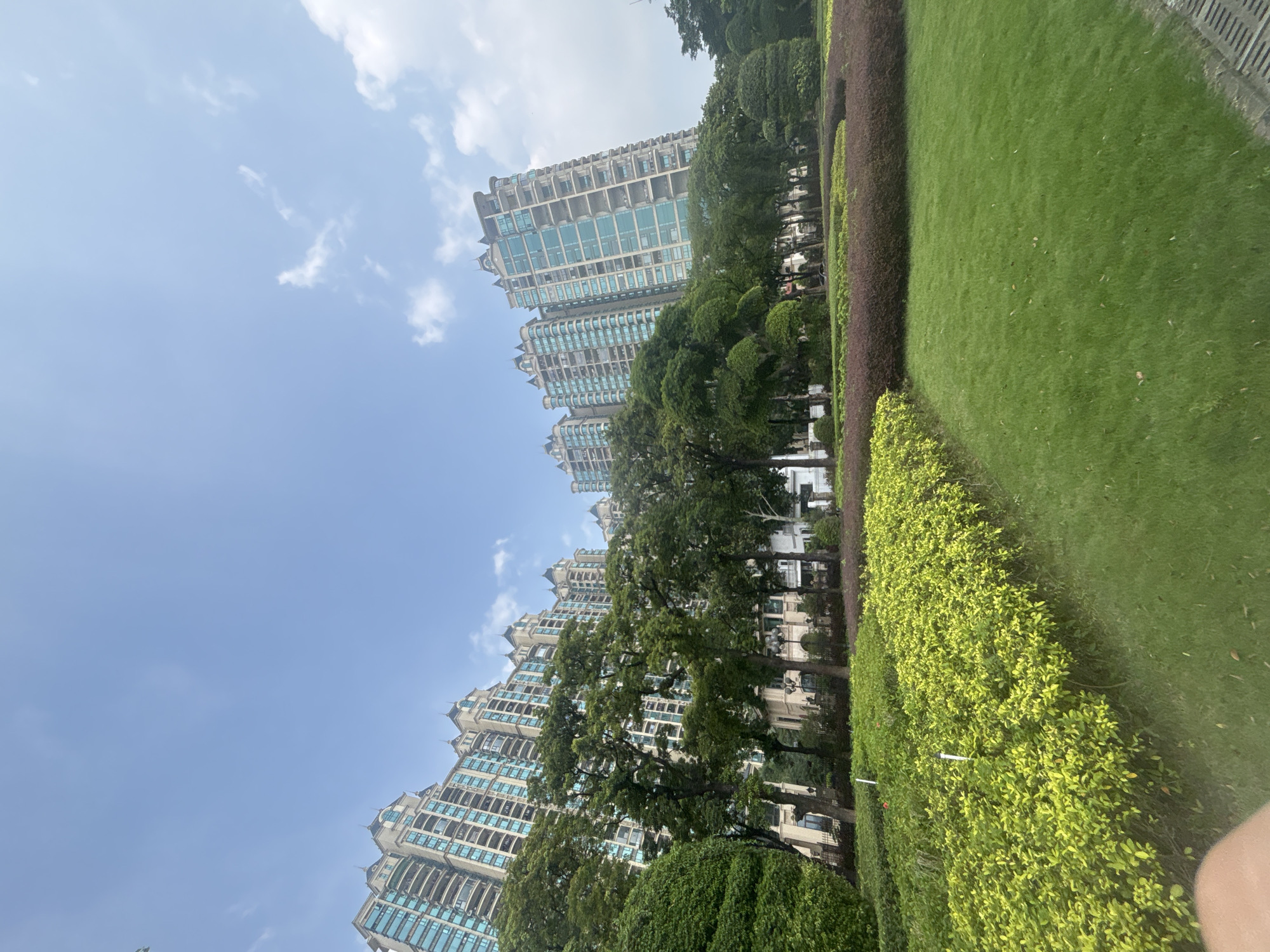 In Nanhai District, China, a wide-angle shot captures a view of modern residential buildings with light-colored facades and blue-tinted windows, interspersed with lush greenery. In the foreground, a neatly manicured lawn leads to a row of vibrant green bushes, followed by a darker, mulched garden bed with some purple-hued plants. Behind the gardens, mature trees with dense foliage stand, and further back, the multi-story apartment buildings rise against a clear blue sky with scattered white clouds. The architectural style of the buildings suggests a contemporary urban setting, likely a residential complex. The scene is devoid of people, focusing on the urban landscape and its manicured green spaces.
