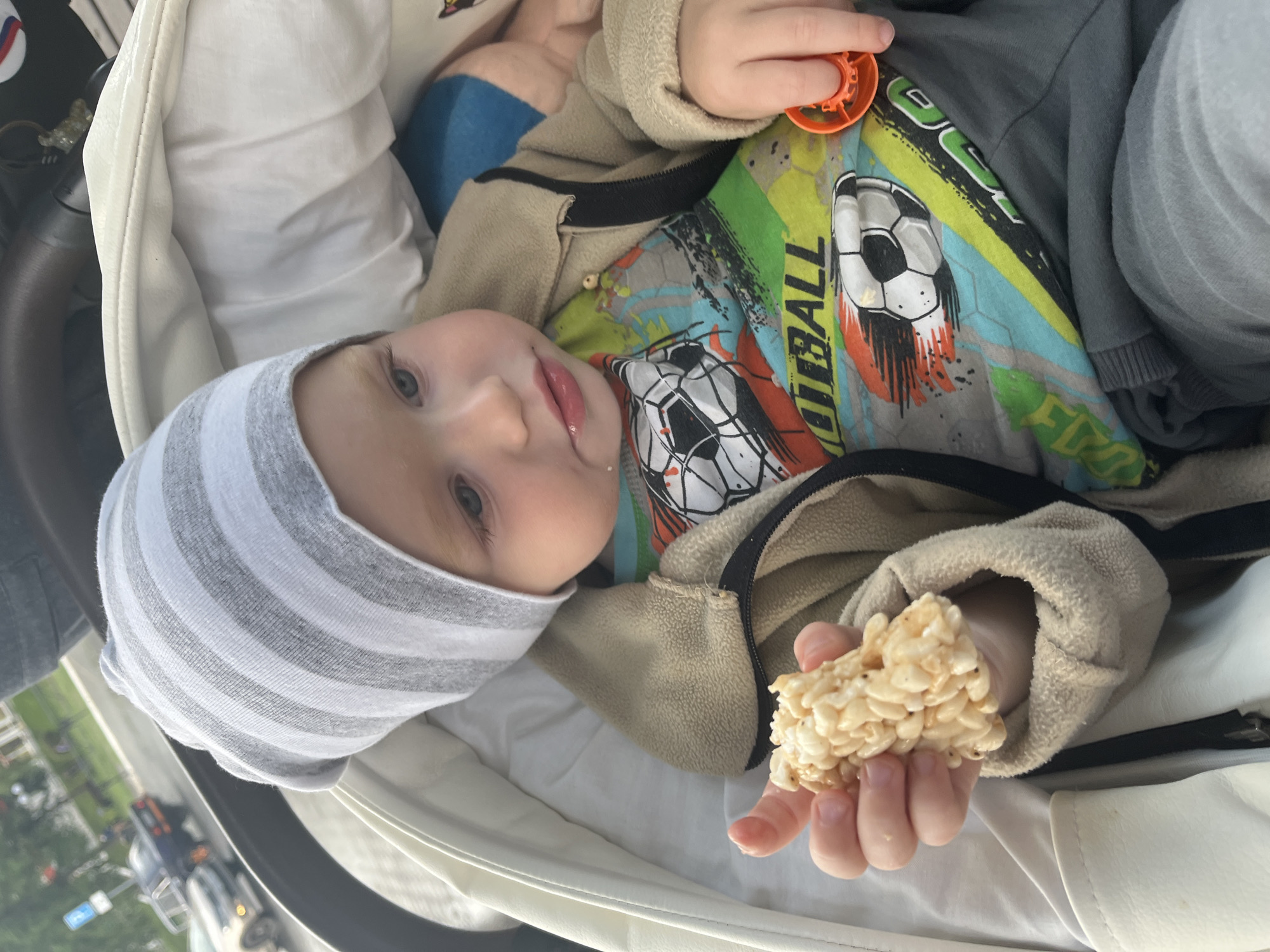 In Chaikovsky, Russia, a close-up shot captures a toddler wearing a gray and white striped beanie and a tan jacket over a colorful t-shirt with soccer ball graphics. The child is seated in a stroller, holding a rice crispy treat in their right hand and an orange toy in their left. The background shows a blurry urban scene with cars and trees, suggesting an outdoor setting. The toddler has light skin and blue eyes, with a piece of food residue visible on their chin. The lighting appears to be natural daylight.