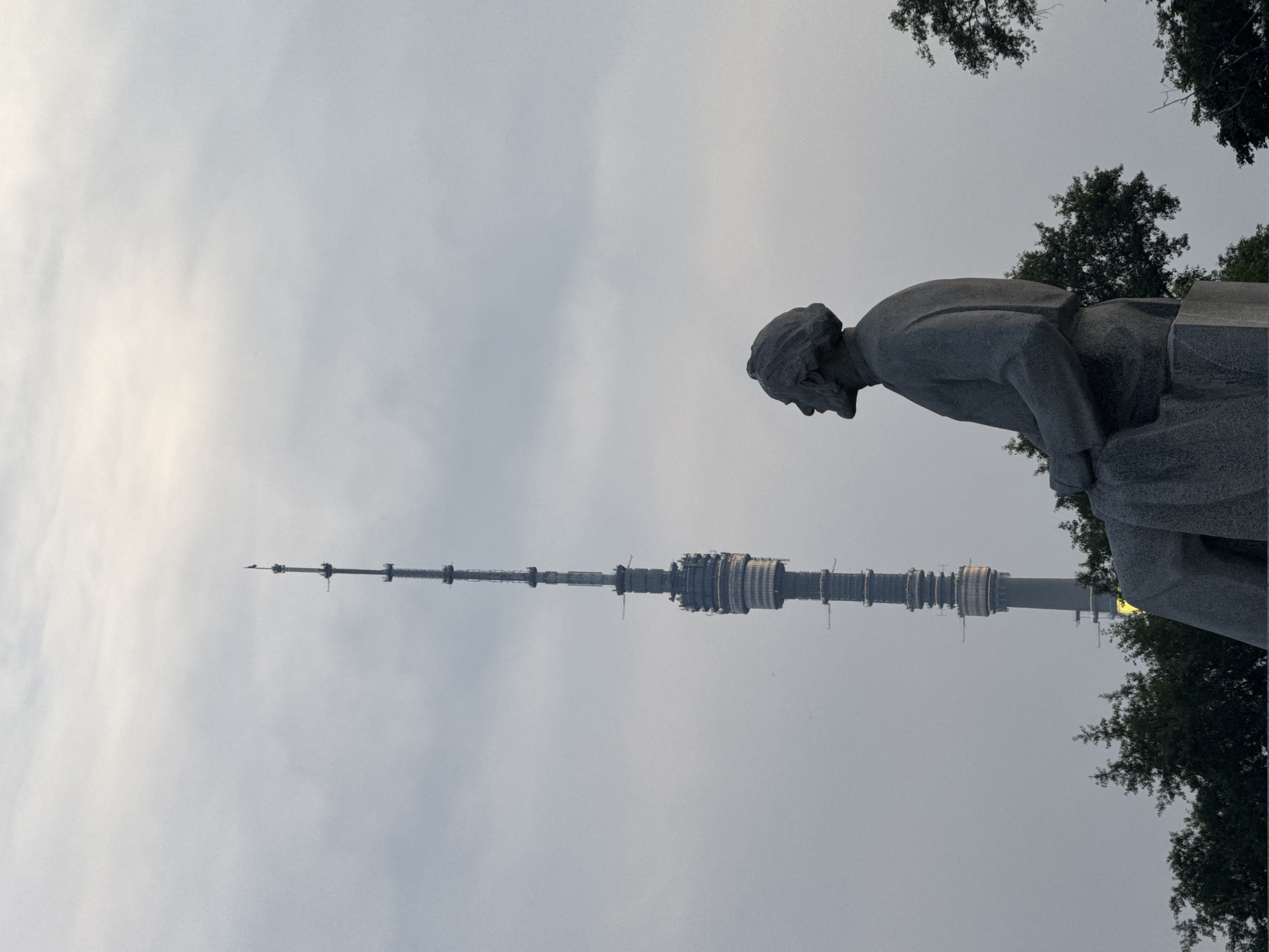 The image captures a view of the Ostankino Tower in Moscow, Russia, seen from behind a large stone statue of a man. The statue is dark gray and depicts the figure seated, with his head turned to the left, looking upwards towards the tower. He has a beard and is wearing clothing that drapes around him. The Ostankino Tower, a prominent landmark, stands tall in the background against a cloudy sky, with its complex antenna structure clearly visible. The tower appears to have lights on in its lower sections. In the foreground and along the bottom of the frame, there are dark green trees. The overall atmosphere is somber due to the overcast sky and the muted colors.