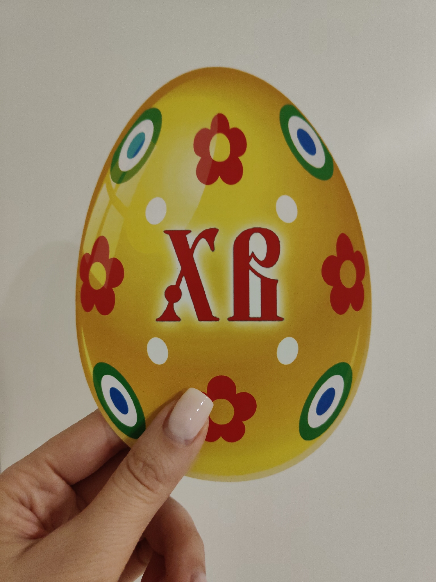 A hand is holding a yellow Easter egg decoration. The egg is adorned with red flowers, white dots, and circular patterns with blue, white, and green rings. The Greek letters "XP" are prominently displayed in red on the egg. The background is a plain, light-colored surface.