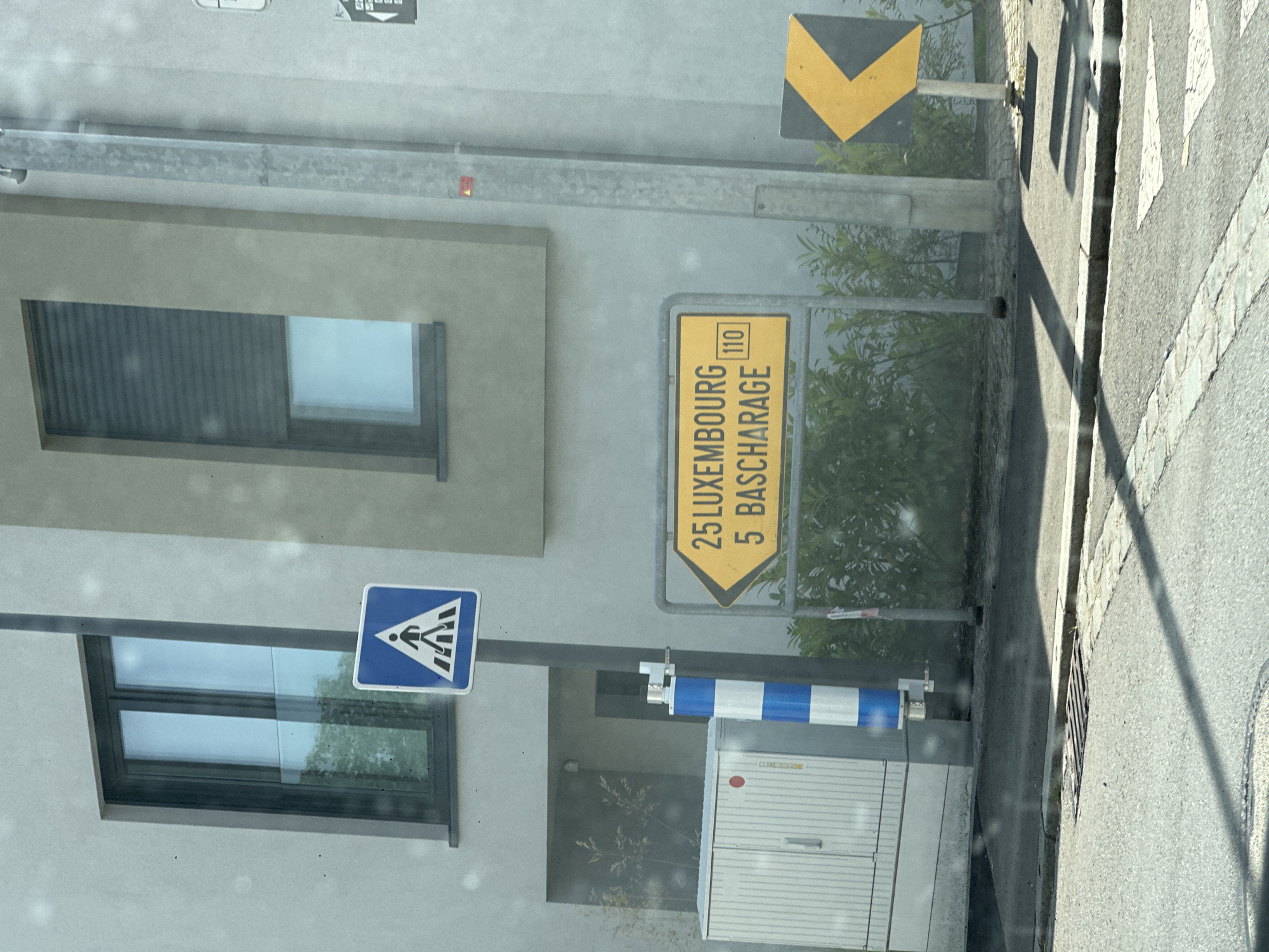 The image captures a street scene in Käerjeng, Luxembourg. A yellow directional sign is prominently displayed, indicating "25 LUXEMBOURG" and "5 BASCHARAGE" with a speed limit of "110". To the left of the directional sign, a blue and white striped pole is visible. Further left, a white utility box with vertical ribbed siding and two doors is situated. Overhead, a pedestrian crossing sign, a blue square with a white silhouette of a person walking on a crosswalk, is mounted on a pole. The background features a modern building with light-colored walls and two windows, one with dark shutters and the other with clear glass. A dark-colored chevron sign pointing left is visible on the right side of the image. The ground is paved with asphalt, and a concrete curb with cobblestone detailing is present. Green foliage and trees are visible behind the signs and utility box, suggesting an outdoor, possibly suburban or urban, setting. The lighting indicates it is daytime, with shadows cast on the road.