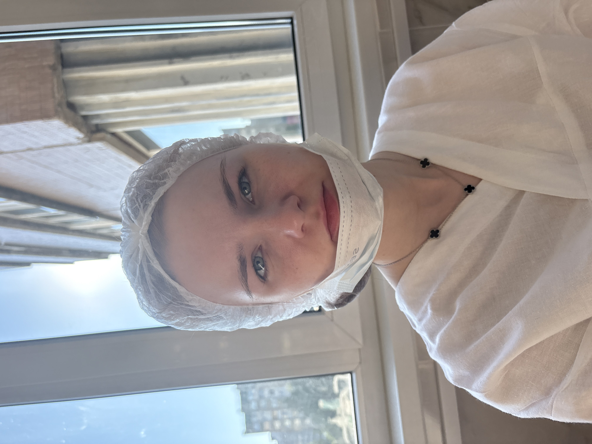 The image is a portrait of a young woman taken indoors, positioned in front of a large window. She is looking directly at the camera with a neutral expression. She has light-colored eyes, possibly blue or green. Her head is covered with a disposable white hair net, and a light blue medical mask is pulled down below her chin, revealing her nose and mouth. Faint text, resembling "CE", is visible on the mask.
She is wearing a white, seemingly loose-fitting garment, possibly a medical gown or smock, which covers her upper body. A delicate necklace with two small black clover-shaped charms is visible around her neck, resting on her collarbone.
The background consists of the window view, showcasing natural daylight. The upper portion of the outside view reveals architectural elements, including what appears to be corrugated metal and brick structures of buildings, suggesting an urban or industrial setting. The lower part of the window shows a clear blue sky and more distant buildings, indicating an outdoor landscape. The bright illumination suggests the photo was taken during the daytime. The overall scene implies a professional environment, such as a clinic or beauty salon.