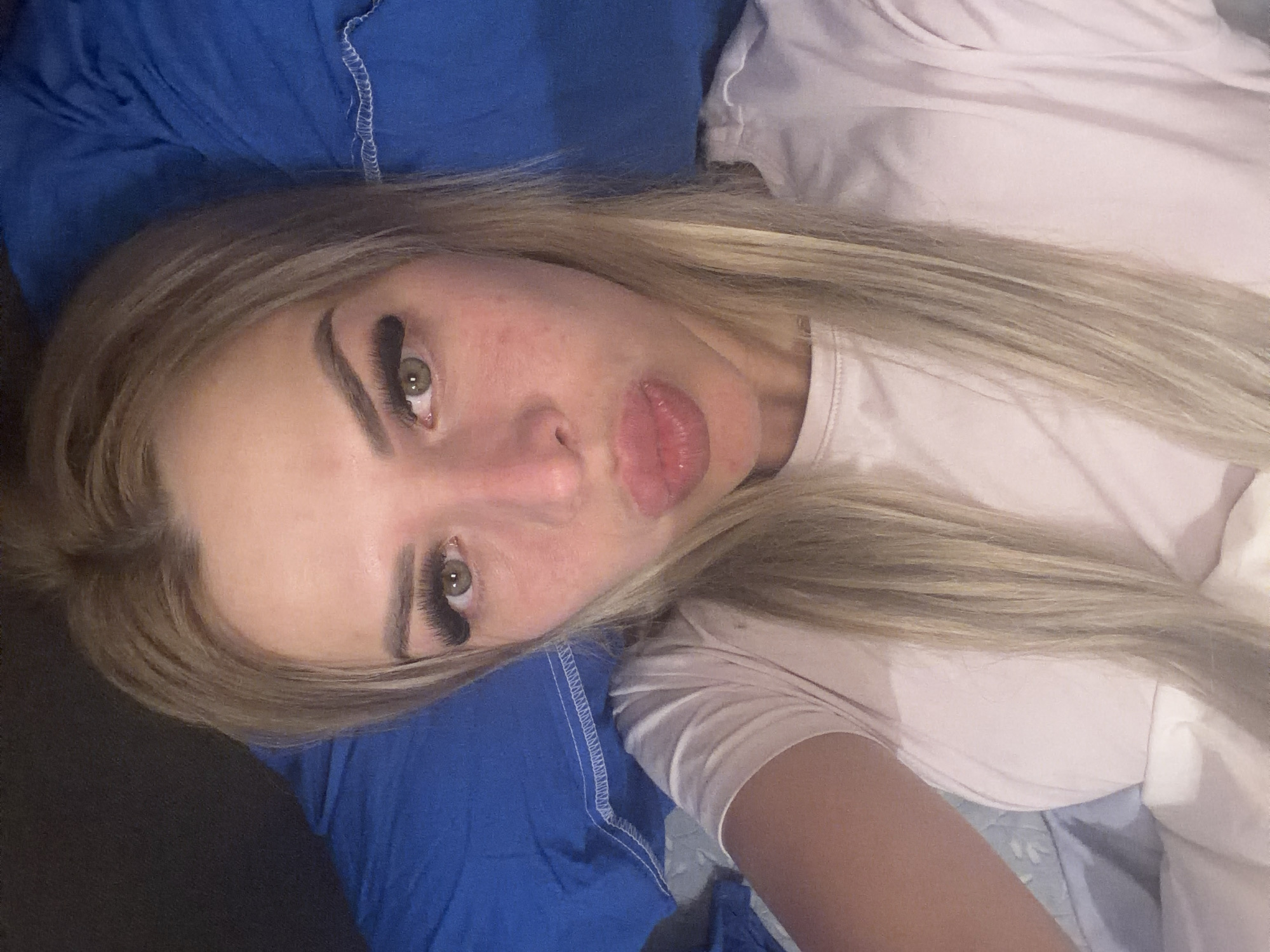 An overhead, close-up shot shows a blonde woman lying on a bed. She is wearing a light pink t-shirt and has dramatic false eyelashes. Her lips are pursed, and she is looking directly at the camera. The background consists of a blue comforter and pillow. There are no discernible landmarks or indicators of the specific location in Kyiv, Ukraine. The lighting appears to be artificial and warm, suggesting an indoor setting, possibly a bedroom.
