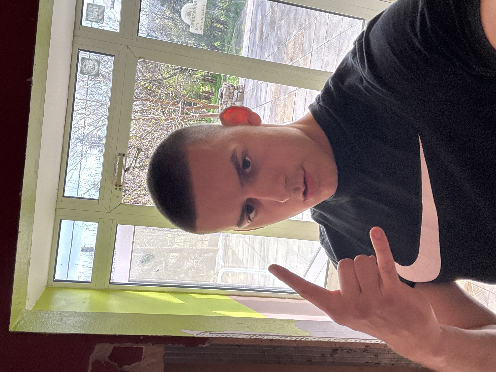 The image, rotated 90 degrees counter-clockwise, shows a young person indoors, posing near a window. The subject, a male with a buzz cut and a piercing on his left ear, wears a black t-shirt with a prominent white Nike swoosh logo. He looks directly at the camera with a neutral expression, holding his right hand up in a "shaka" or "hang loose" gesture.
The interior setting features a window with a light green sill and frame. The wall below the sill is a dark red/brown, showing significant peeling paint and exposed underlayers. Through the multi-pane window, an outdoor scene is visible. It includes a paved area, likely a patio or courtyard, and trees with bare branches and some budding green leaves, suggesting springtime. The natural lighting indicates daytime, possibly an overcast or partly cloudy day.
A mirrored security sticker is affixed to an upper window pane, displaying text that includes "24 HOUR SECURITY", "RAMS", "REGISTERED", and phone numbers "0800 323232" and "015332", along with a circular emblem. The scene is located in Goritsa, Bulgaria.