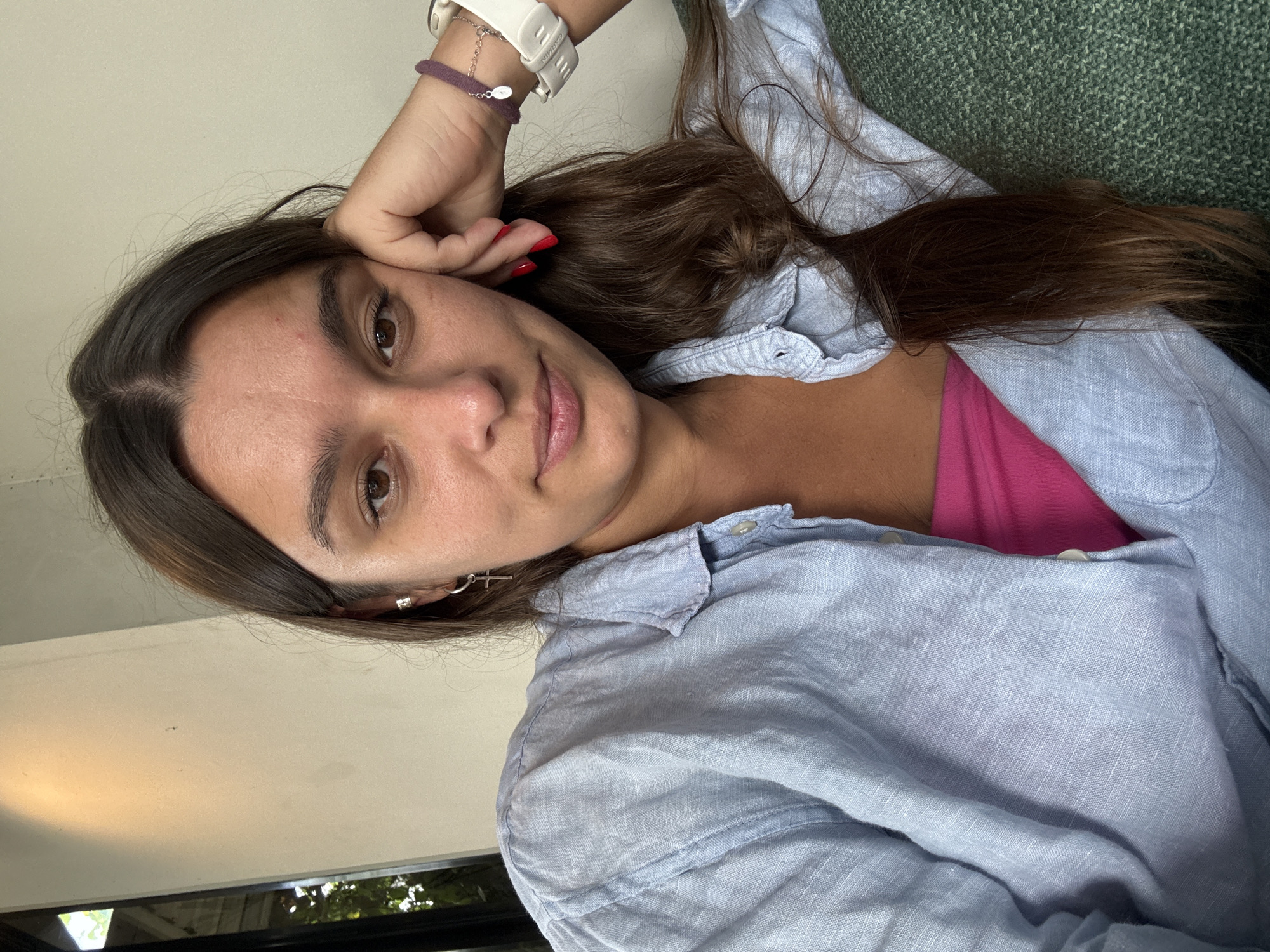 A woman is lying down, possibly on a couch or bed. She has brown hair and is wearing a light blue button-down shirt over a pink top. She is wearing a watch and a bracelet on her left wrist. Her left hand is resting on her forehead. Her right arm is to the side. The photo was taken indoors. The location is not clear from the image and can't be confirmed from Ubud District, Indonesia.