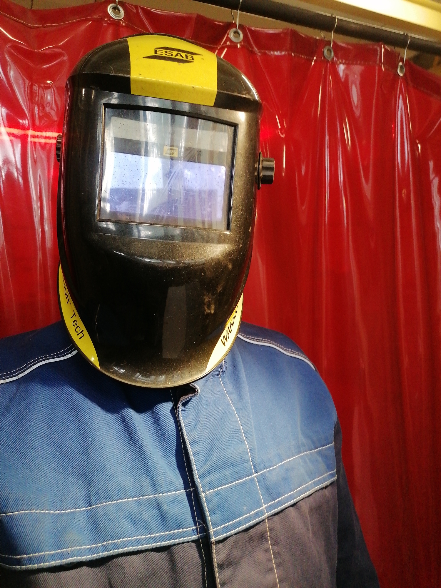 A welding helmet sits on a blue and gray work shirt in front of a red industrial curtain in an unknown location. The helmet is black with a yellow stripe across the top and the brand name "ESAB" printed on it. Yellow lettering on the lower part of the helmet reads "WARRIOR Tech". The curtain is hung from a metal bar with metal hooks.