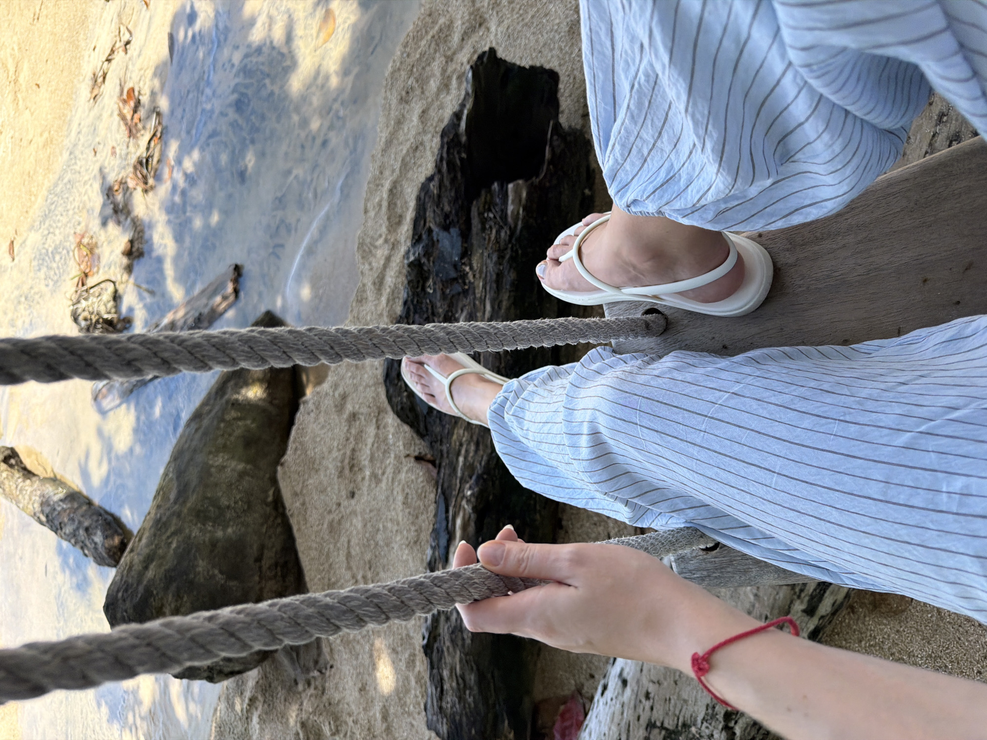 A first-person perspective shot shows a person sitting on a wooden swing suspended by thick ropes. The person is wearing light blue striped pants and white flip-flops. One hand is visible, holding onto one of the ropes and wearing a thin red string bracelet. The swing is positioned over a sandy beach area with driftwood logs scattered around. In the background, shallow, clear water laps against the shore, with more logs submerged or partially visible in the water. The scene appears to be a serene, natural outdoor environment.