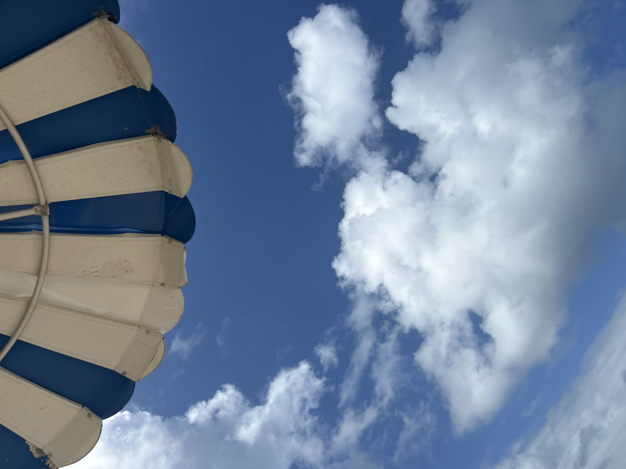 An upward-angled shot features a blue and white striped sunshade to the left, contrasted with a blue sky dotted with white cumulus clouds. The sunshade's fabric panels are visibly worn. The environment consists of fair weather conditions and a seaside location, which suggests a possible place in Le Gosier, France. There are no people visible in the image. The key objects are the sunshade and the clouds in the sky.