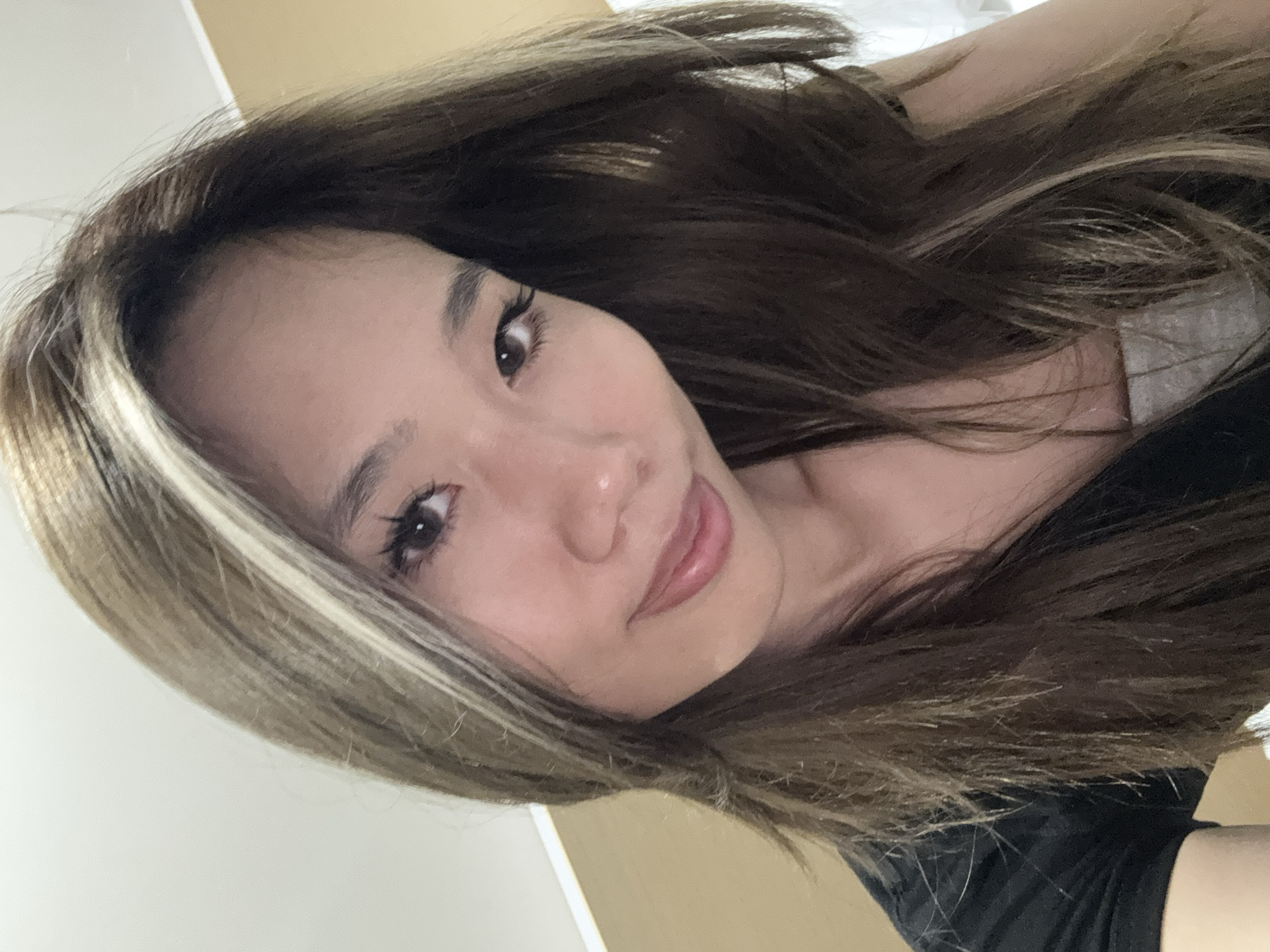 A close-up selfie of a woman with long brown hair highlighted with blonde streaks. She has fair skin, dark eyes, and is smiling subtly. She is wearing a black shirt. The background is a blurred, light-colored wall. The location is not evident and there is no discernible information to indicate that the scene is in the Yuzhong District, China.
