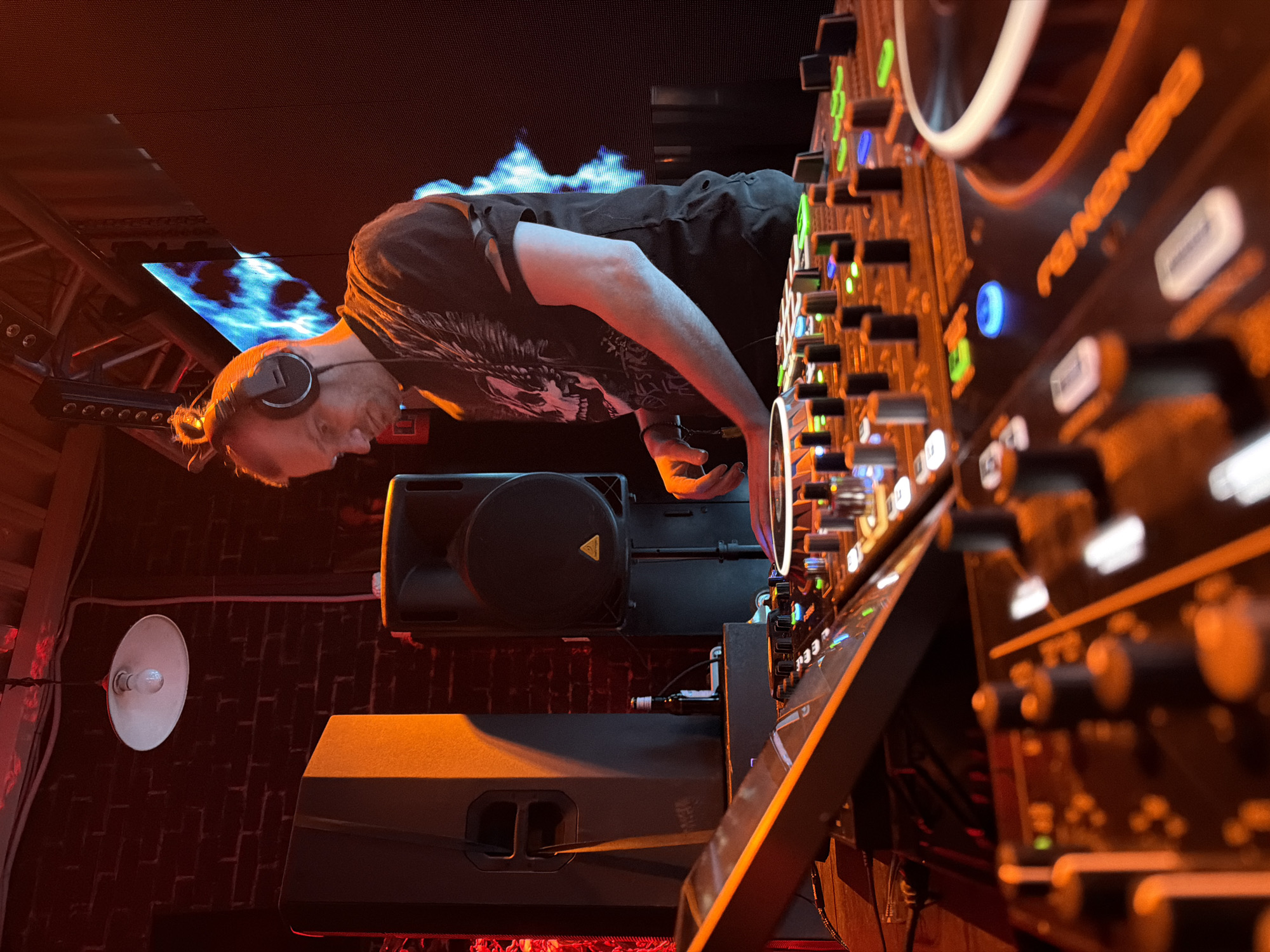 A DJ is playing music at what appears to be a club or event in Votkinsk, Russia. He is wearing headphones and leaning over a Pioneer DJ mixer, likely adjusting settings or mixing tracks. The environment features exposed brick, speakers, a lighting fixture, and a screen displaying blue visuals, creating a warm ambiance.