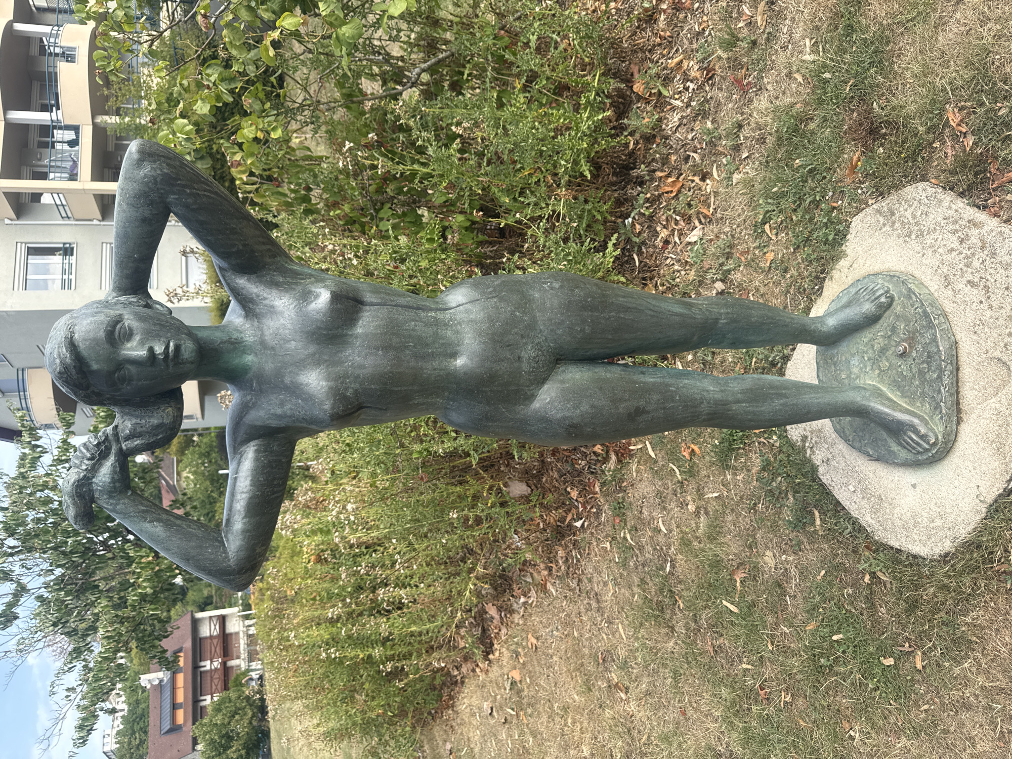 This is a full shot of a bronze statue of a female figure standing on a circular stone base. The statue is located outdoors in a park-like setting. The figure is depicted with her arms raised, holding something in her hands near her head, possibly adjusting her hair. She is naked and slender. Behind the statue, there is a dense green bush and a patch of dry grass with fallen leaves. In the background, there are modern apartment buildings with balconies and windows, as well as some trees and a hillside with houses. The sky is partly cloudy. The statue is positioned in a slight upward angle from the viewer.