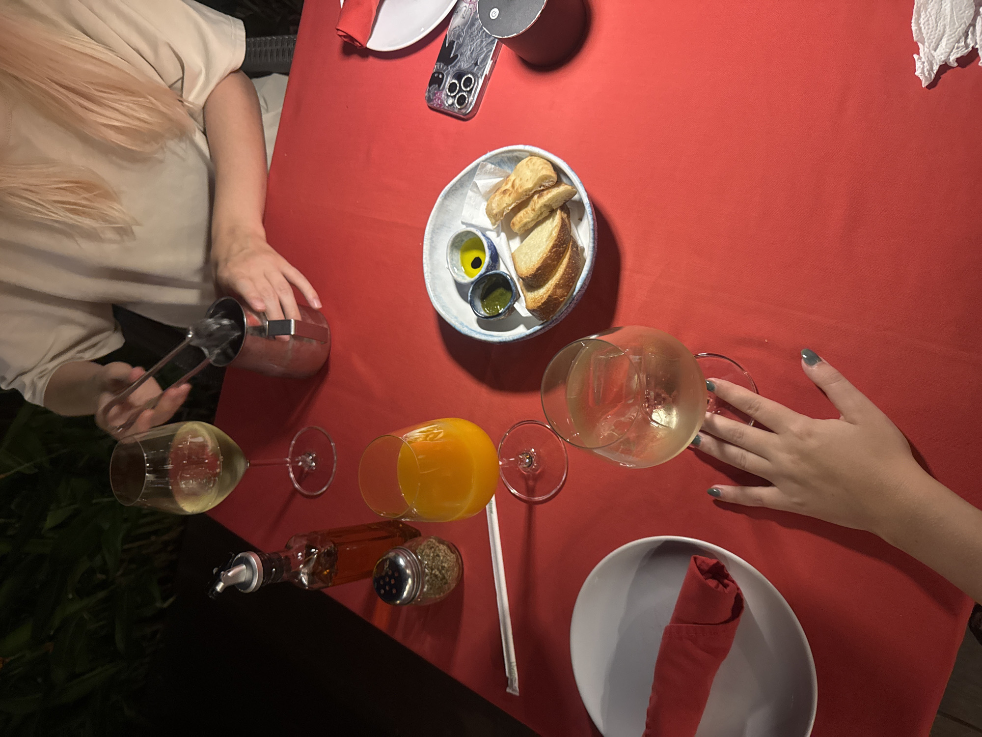 An overhead shot captures a vibrant red tablecloth set for a meal outdoors, likely in Chiang Mai City Municipality, Thailand, given the lush dark foliage visible in the background. Two wine glasses, one filled with white wine and the other with ice water, are prominent. A glass of orange juice sits nearby, along with a bottle of olive oil and a shaker of dried herbs. A small bowl contains slices of toasted bread, served with two dipping sauces, one olive oil and balsamic vinegar, the other a green herb-based sauce. To the right, a smartphone with a decorative case rests on the table, next to a small, dark cylindrical object. A person's hand with dark green nail polish is reaching towards the ice water glass, suggesting an interaction with the scene. The lighting is dim, indicating an evening setting, and the overall atmosphere is relaxed and inviting.