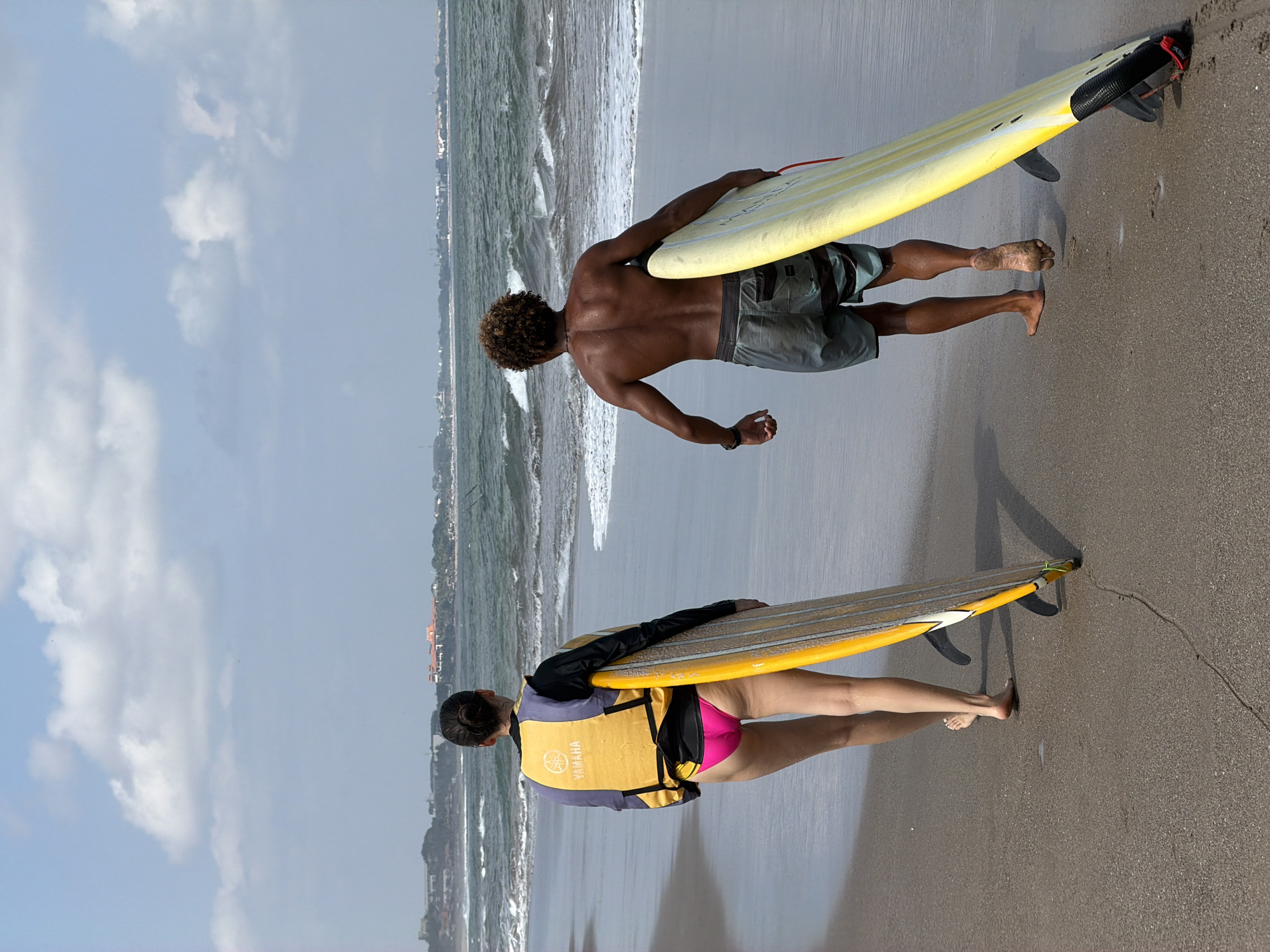 Two individuals, one female and one male, walk along the shore of a beach in North Kuta, Indonesia, each carrying a surfboard. The woman, viewed from behind, wears a yellow life vest over a black rash guard and bright pink bikini bottoms. She holds a long, yellow surfboard with a wooden-grain pattern vertically, its fins visible. The man, also viewed from behind, has curly hair and wears grey and blue patterned swim shorts. He carries a long, yellow surfboard, also vertically, with red leash visible near the bottom. The ocean is visible in the background with moderate waves and white foam breaking on the shore. The beach is sandy, and a faint line of buildings can be seen in the distance along the coastline. The sky is partly cloudy with a bright, hazy sun.