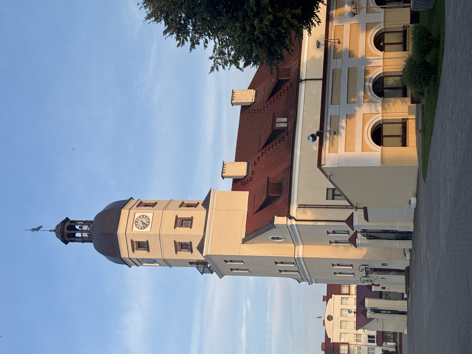 The image shows the Nesvizh Town Hall in Nesvizh, Belarus, under a clear blue sky with scattered clouds. The Nesvizh Town Hall is a prominent building with a distinctive clock tower. The tower is tall and octagonal, topped with a dark spire and a weather vane. The main body of the town hall is a pale yellow color with red-tiled roofs. The building has multiple windows, some of which are arched and decorated with ornate trim. In the foreground, there is a paved square or plaza, and to the right, a lush green tree. The lighting suggests it is either early morning or late afternoon, with the sun casting long shadows. In the background, other buildings of similar architectural style can be seen.