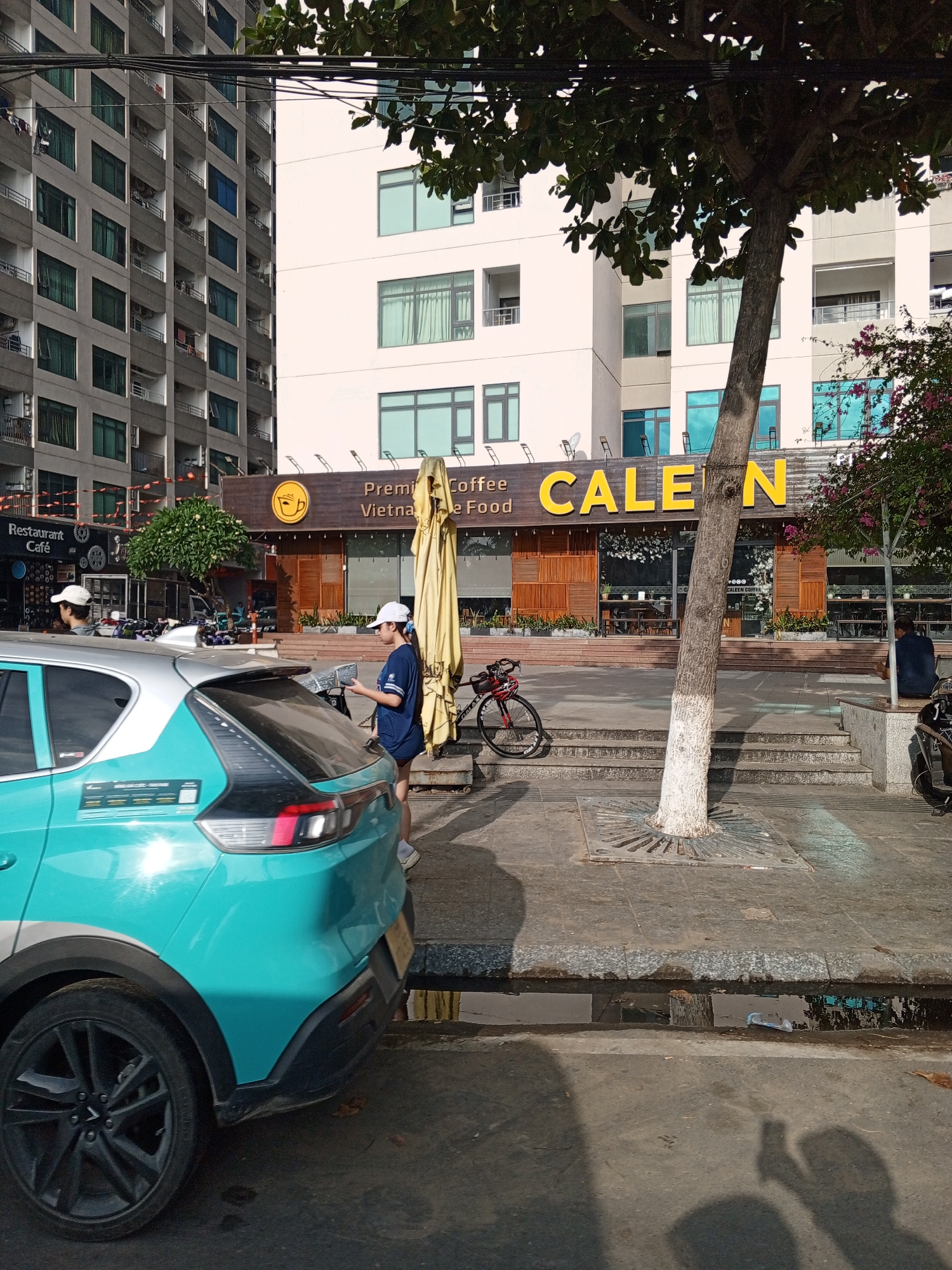 The image depicts a street scene in Bac Nha Trang Ward, Vietnam. A teal and silver car is parked on the right side of the frame. In front of the car, a person wearing a dark blue shirt and a white cap is standing next to a large, yellow, folded umbrella. Further to the right, a bicycle is parked. A tree with a white-painted trunk is visible in the foreground, casting a shadow on the pavement. Behind the tree, a building with large windows and wooden paneling is adorned with the sign "CALEN COFFEE FOOD." The building's facade features a large yellow logo with a coffee cup symbol. Several tables and chairs are visible through the windows of the coffee shop. In the background, a tall, gray apartment building with numerous balconies and windows dominates the left side of the image. A smaller, white building with similar window designs is situated to the right of the apartment building and behind the "CALEN COFFEE FOOD" establishment. The ground is paved with concrete and stones, and a small puddle of water is present near the curb.