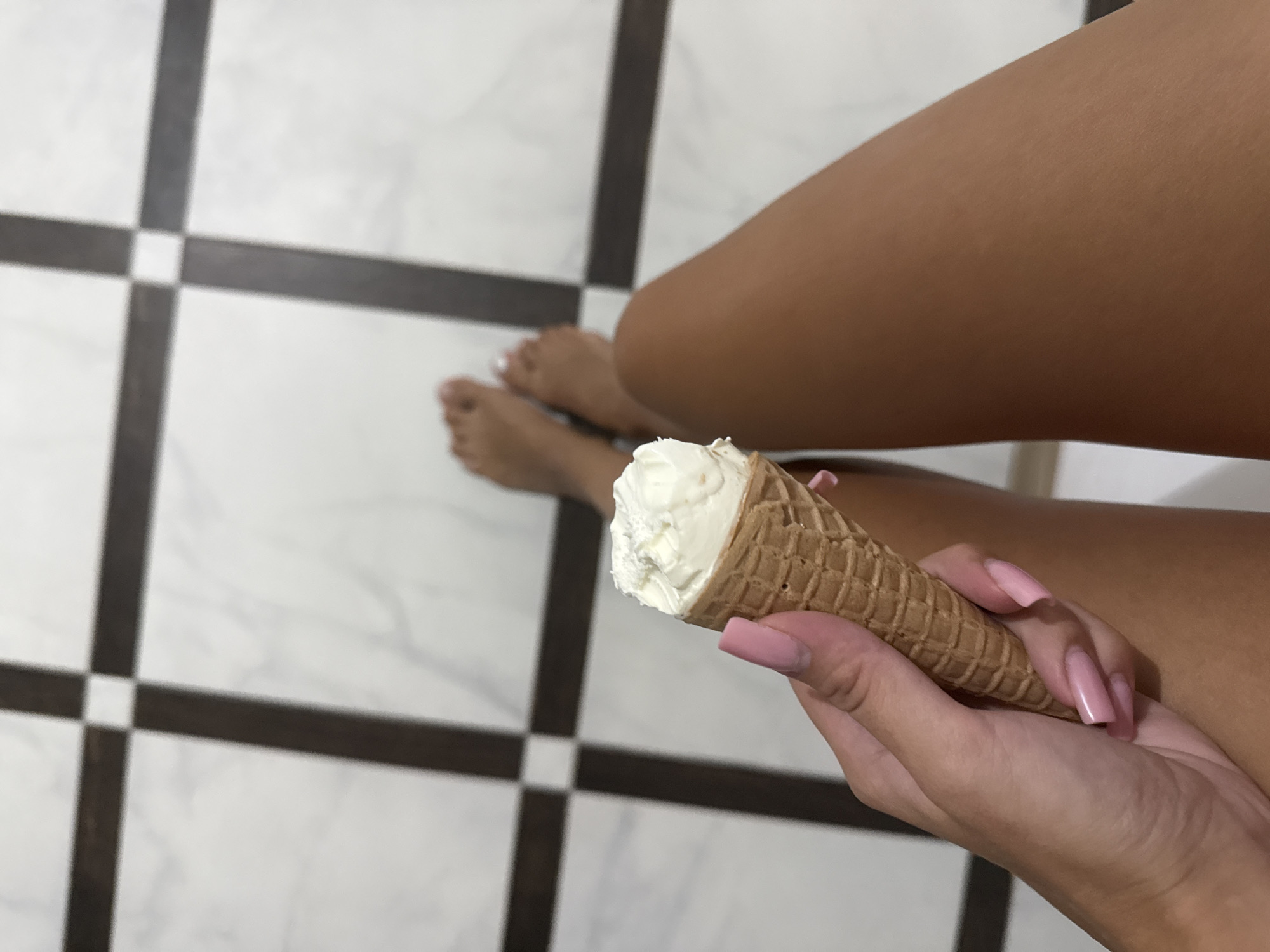 The media file shows a close-up view of a person's hand holding an ice cream cone. The ice cream appears to be vanilla-flavored. The person's fingernails are painted a light pink color. In the background, there are a person's legs and feet, suggesting they are sitting on a tiled floor. The floor has a pattern of white and dark brown tiles, arranged in a grid. The lighting suggests it could be daytime, possibly indoors in a well-lit room or outdoors in bright conditions. The overall mood is casual and relaxed. There is no visible text in the image.