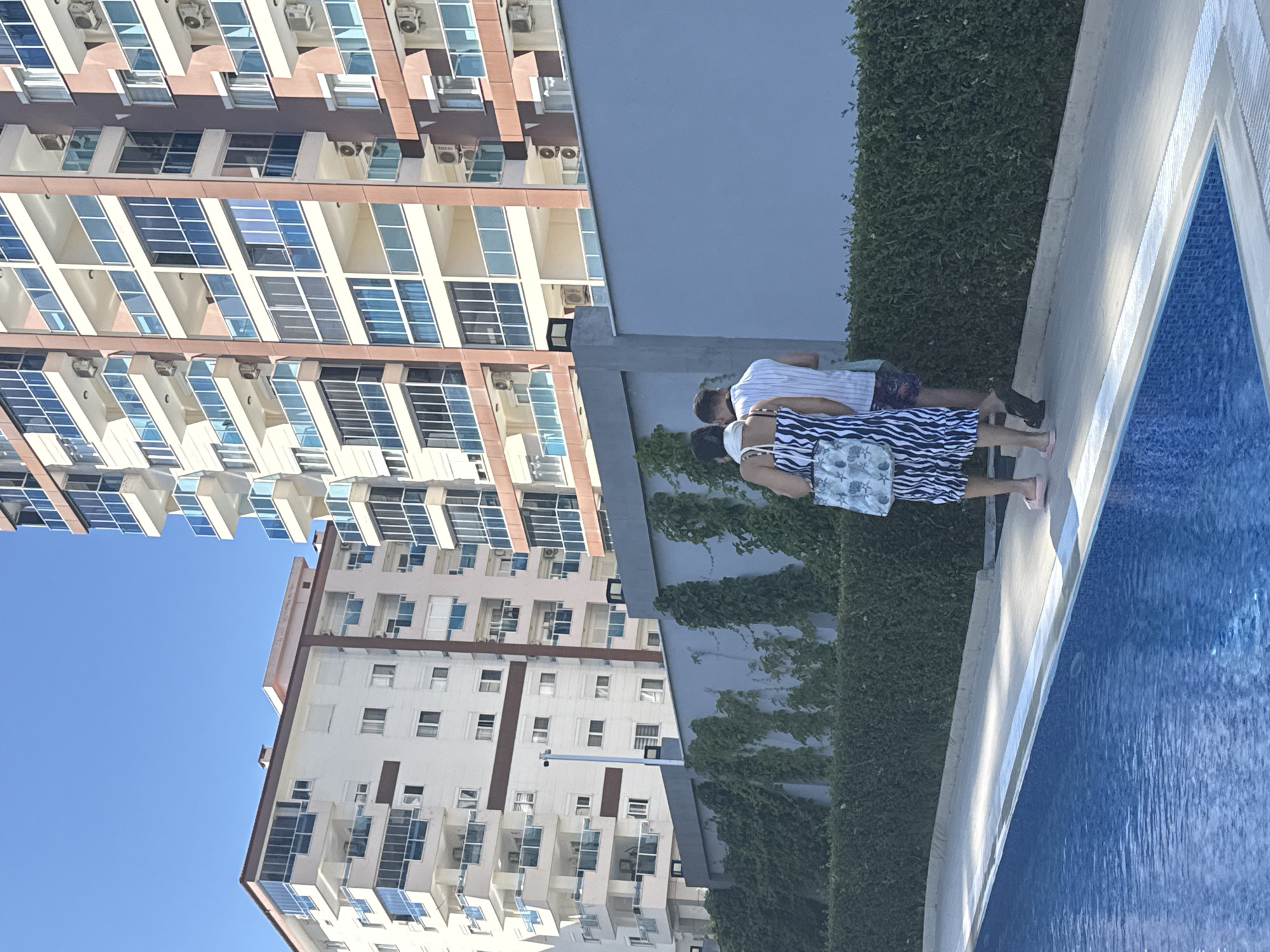 The image captures an outdoor scene. Two people, a man and a woman, stand near a swimming pool. The woman wears a striped dress and carries a bag. The man is wearing a striped shirt and patterned shorts. Beside them is a high green shrub hedge. Behind them, two buildings with multiple windows and balconies rise towards a clear blue sky. The location is in the city of Unknown, Unknown.
